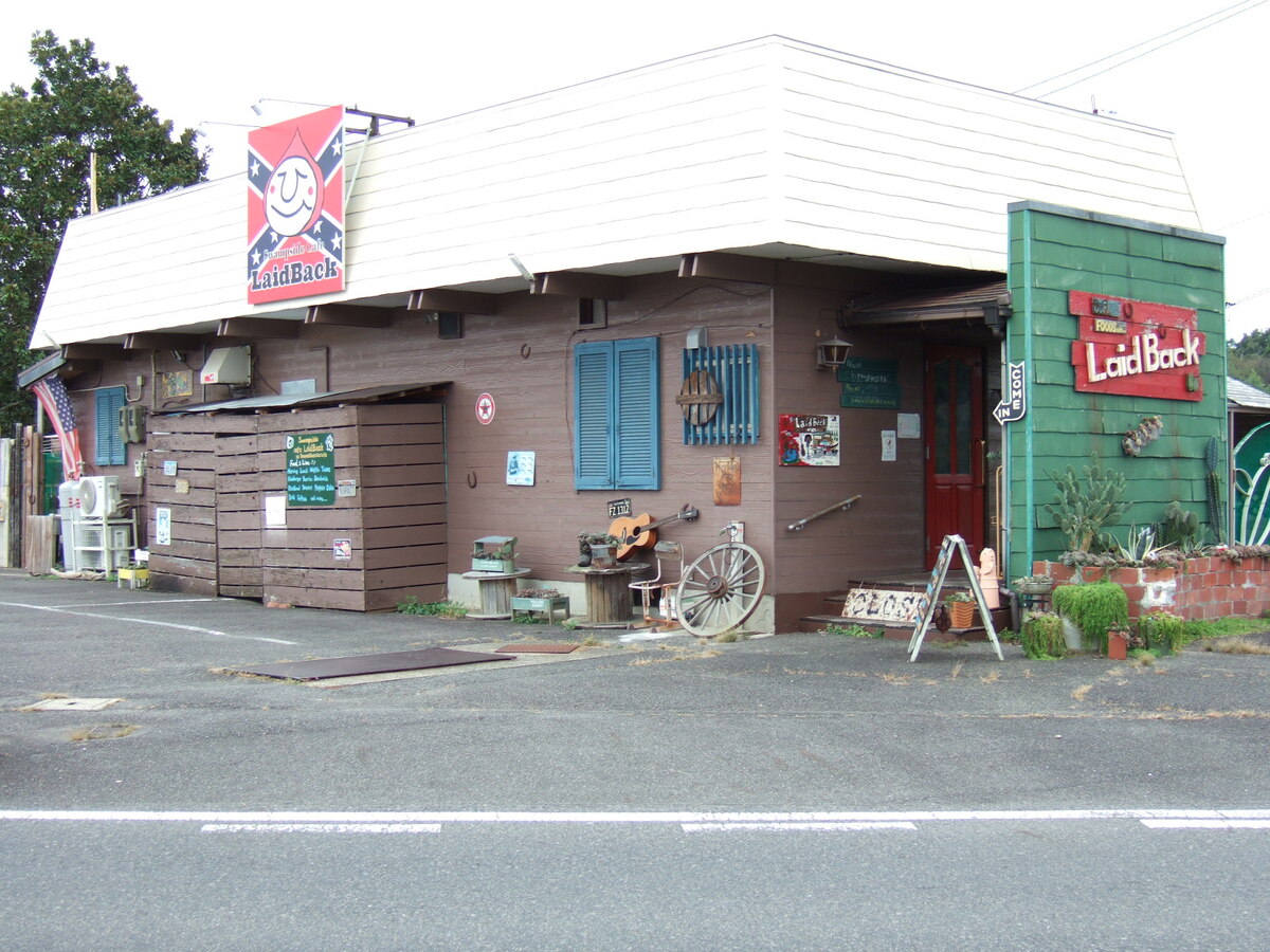 A close-up view of the entrance to the restaurant, a building in the style of a fast-food restaurant, with clapboard siding, decorated with two Confederate flags, one of which has a large white smiley face in the center. A sign on the side of the building reads “LaidBack.” A wagon wheel, a guitar, and American flag, and some other paraphernalia are arranged beside the entrance.