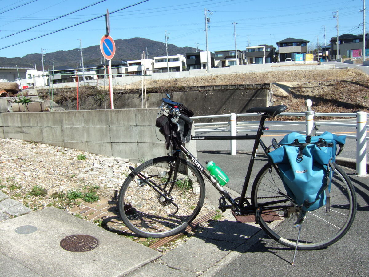 A bicycle with rear panniers parked on the near side of a street bounded by a low concrete wall extended to the right by a guard rail. Beyond the street and above a retaining wall in the distance is a row of half-completed flat-roofed prefab houses.