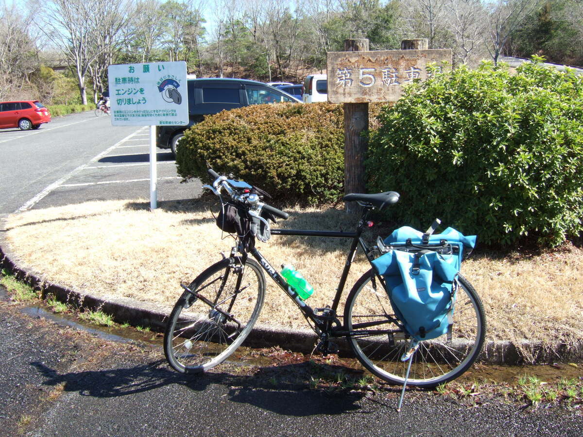 A bicycle with rear panniers parked in front of a traffic island inside a parking lot. A bush grows in the traffic island, and a sign peeks out of the bush reading (in English translation) “Number Five Parking Lot”. To the left of the traffic island a sign in Japanese asks drivers to turn off their engines when parked.