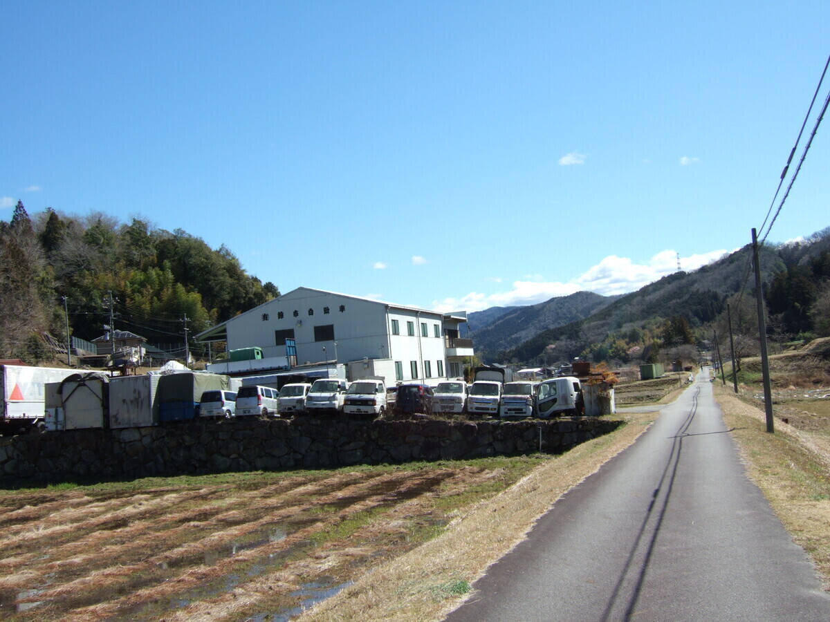 In a rural mountain valley, a road stretches ahead straight as a string. To the left of the road is an industrial lot with a dozen or so out-of-service vans parked in a row next to a large rubbish tip.