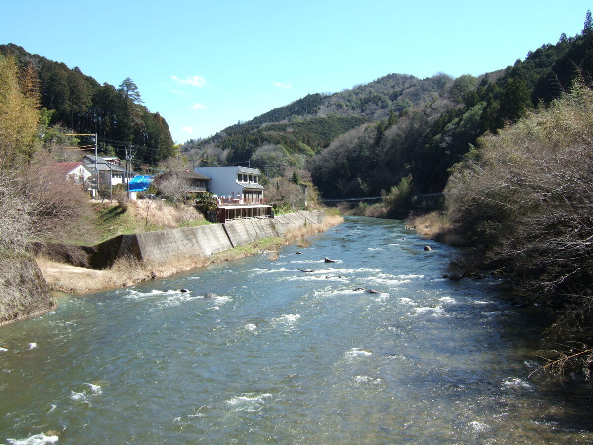 View of a river running through hilly terrain, showing some small whitewater caps. The left bank is shielded by a concrete wall, atop which there isa building that may be a residence or a restaurant with a patio area facing onto the river.