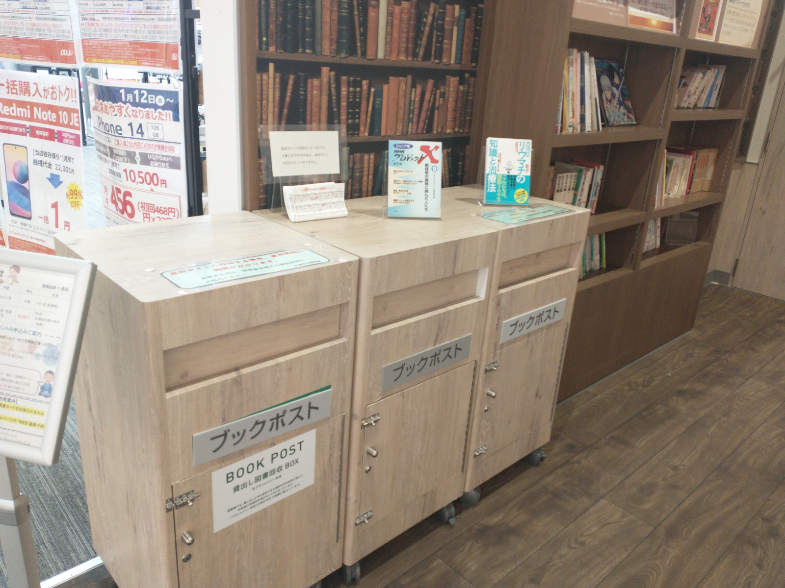 Three “book boxes” for return of borrowed books, standing in a small wood-floored side area of a shopping mall. Behind the boxes is the corner opening into the nook. The mall-side of the corner is covered with advertising flyers for the business next door. The nook side is covered in bookcase wallpaper, but there is an actual bookcase with books deeper in the nook.