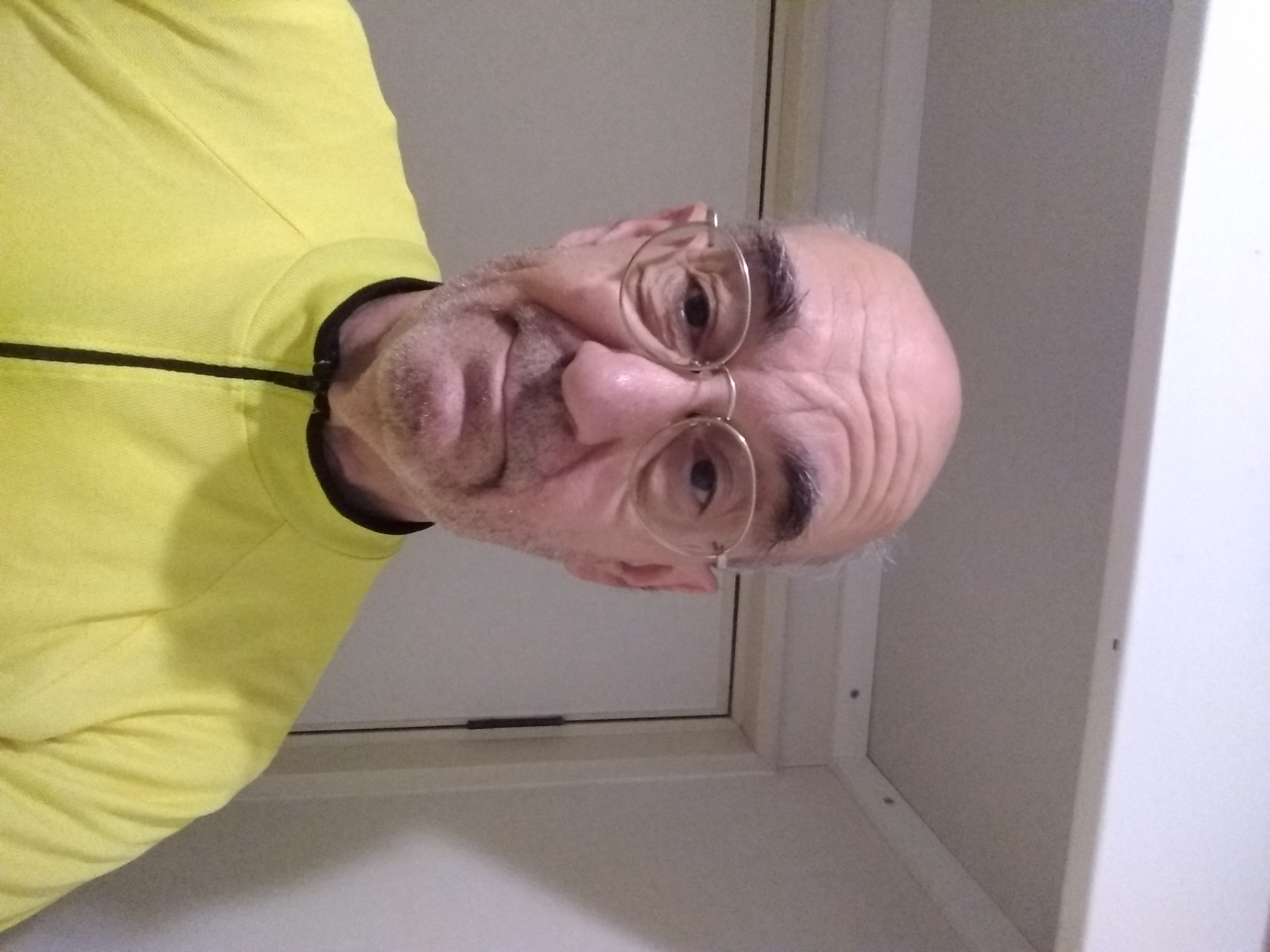 Elderly bald man in a yellow riding jersey and gold-rim glasses smirking at the camera with a weary expression