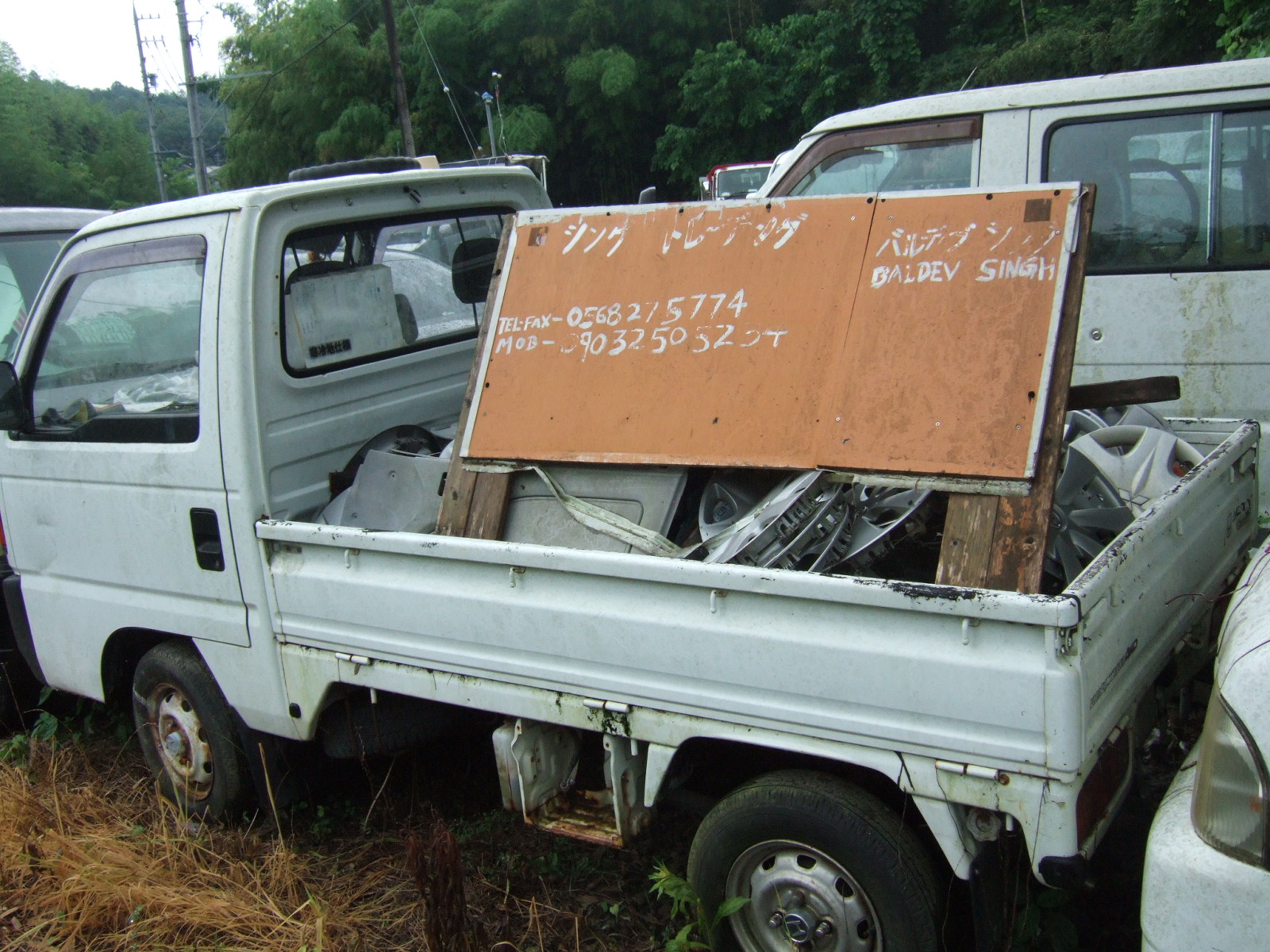 A mini-truck in a lot of used vehicles, with hubcaps and other scrap piled in the bed, and a wooden sign propped up facing the camera, reading (in English translation) “Singh Trading / Baldev Signh” with land-line and cell numbers.