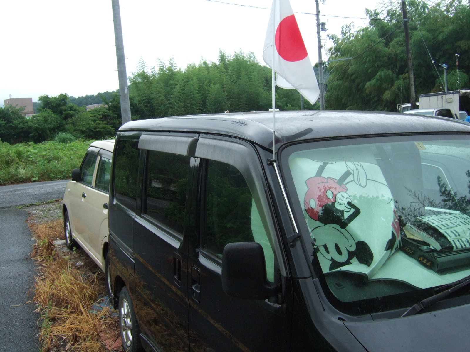 A mini-van in a lot of used vehicles, with a Japanese flag raised on the antenna, and an upside-down image of Mickey Mouse on a sunshade showing through the windshield.