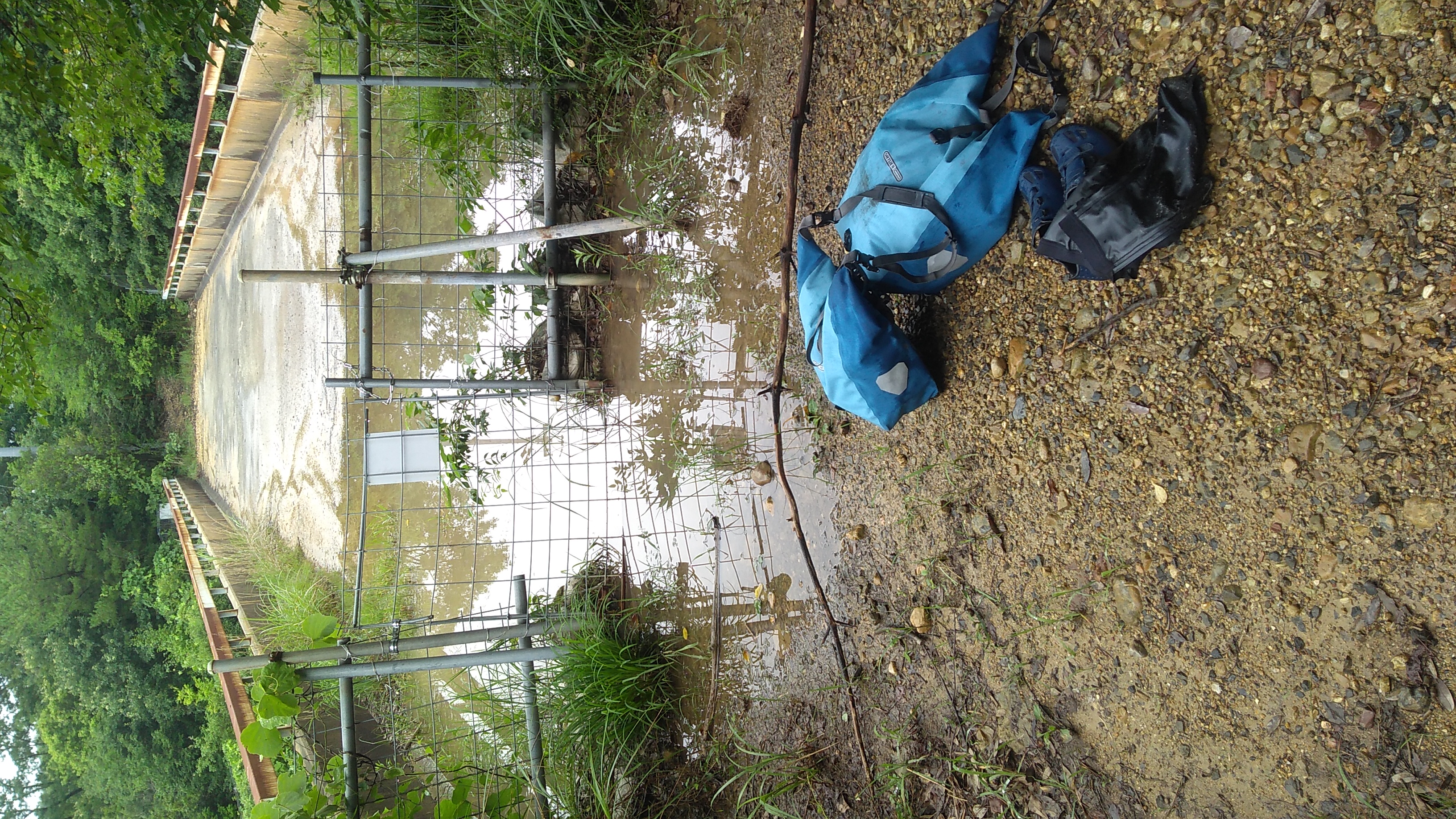 Two panniers and a bag (containing a pair of shoes) on wet ground in front of a fence and gate blocking entry to a long wide steel and concrete footbridge with a puddle covering its near end.