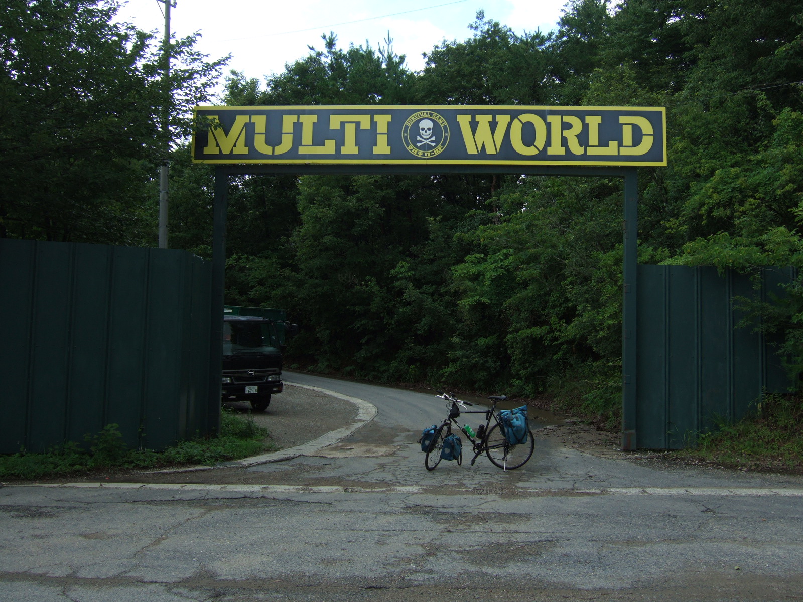 Bicycle with front and rear panniers parked in a broad entrance off a paved street, leading into forestland. A large marquee sign spans the entrance at 3 meters’ height, reading “Multi World,” with a logo between the two words consisting of the words “Survival Game” written in a circle around a skull and crossbones.