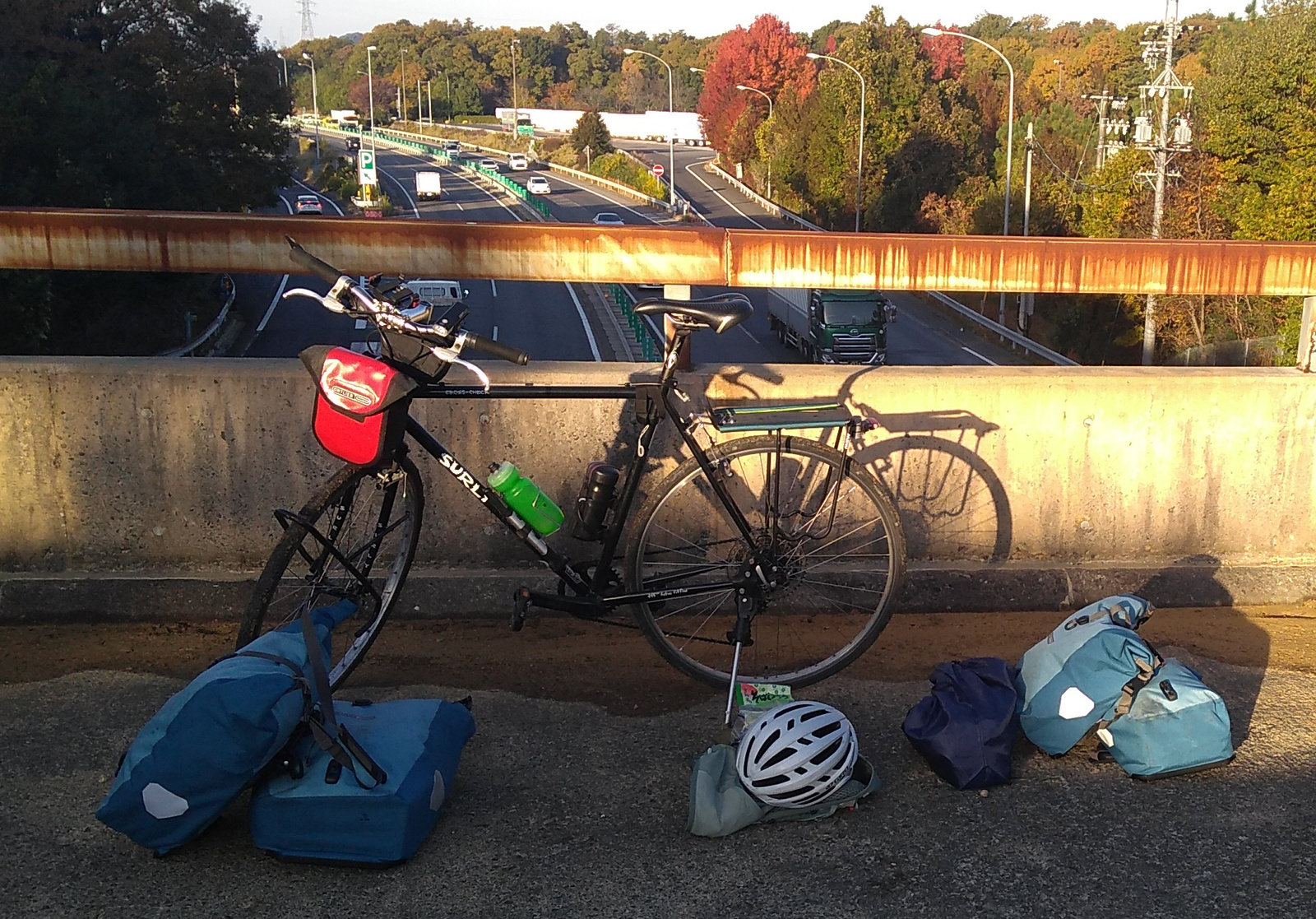 A black gravel bike parked at the rusty railing of a freeway overpass walkway, with four pannier bags and a helmet arranged on the ground in front.