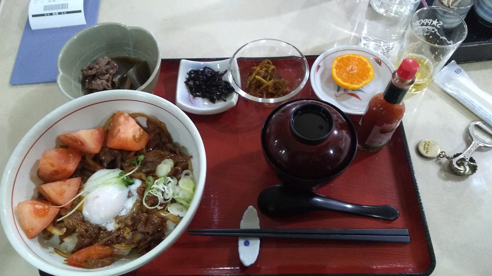 A set meal on a tray, consisting of a bowl of sliced beef braised in a heavy sauce served over rice with sections of fresh tomato and a poached egg on top, portions of pickled lotus root, hijiki, kinpira gobō, orange, and miso soup. A small empty glass of beer stands to the side.