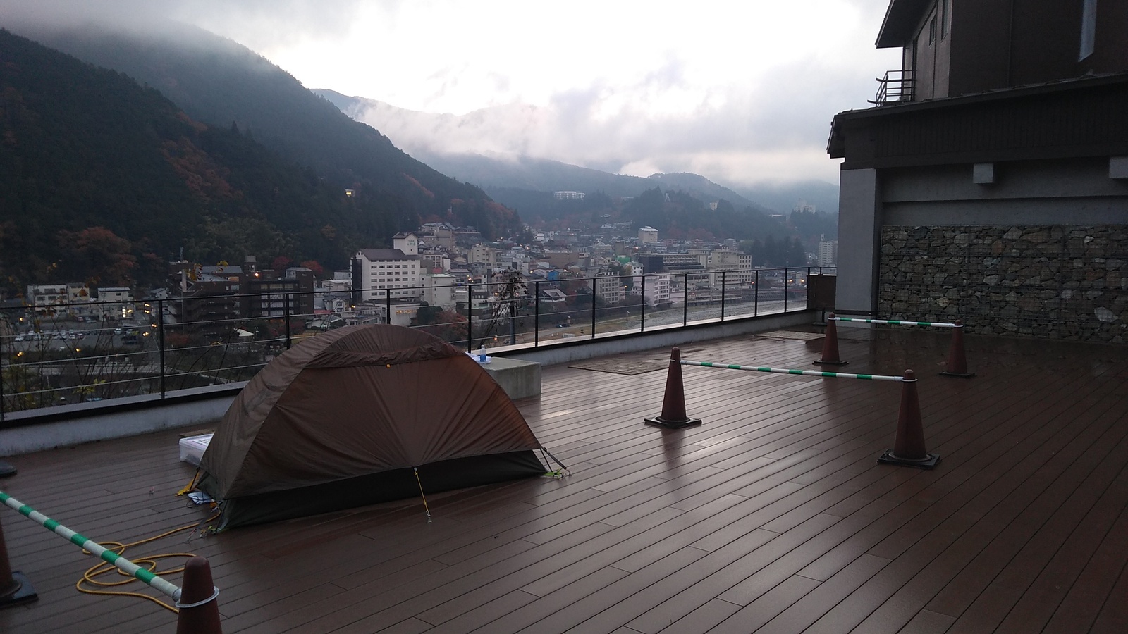 A one-person tent pitched on a rooftop deck, overlooking a town nestled in a mountain valley, partly shrouded in mist with wisps of cloud in a morning sky.