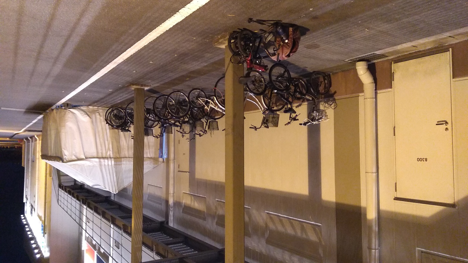 The wall of a two-story utilitarian structure painted beige and bathed in fluorescent light. A row of bicycles is parked along the wall, and a collapsed folding bicycle is locked to a pillar near the wall, with a ratty red backpack on the ground next to it.