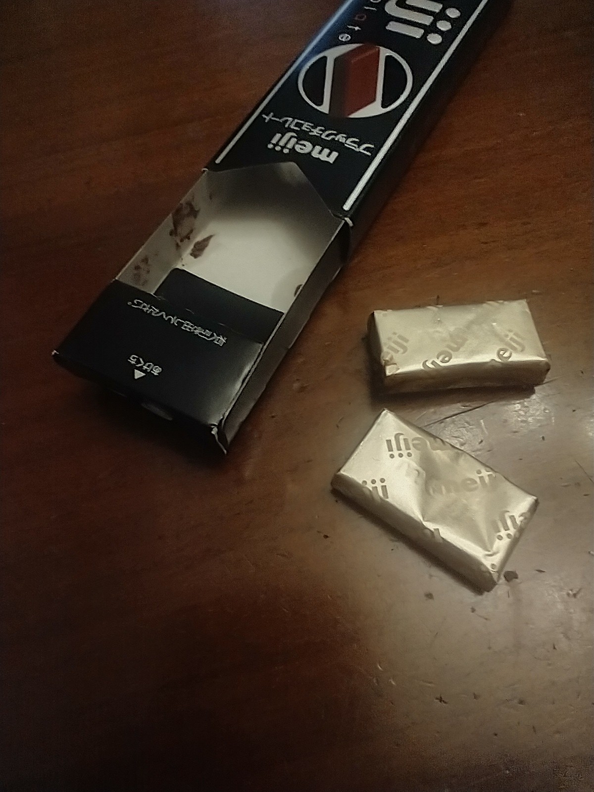 Two wrapped pieces of chocolate beside a Meiji Black Chocolate box, one piece with a dent in its center.