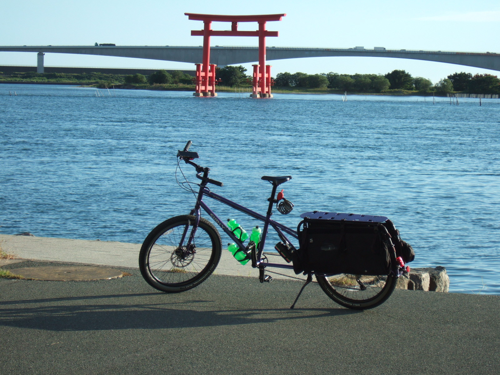 A cargo bicycle parked at water’s edge, with a Japanese shrine gate (“torii”) in the background, and a freeway overpass beyond that.