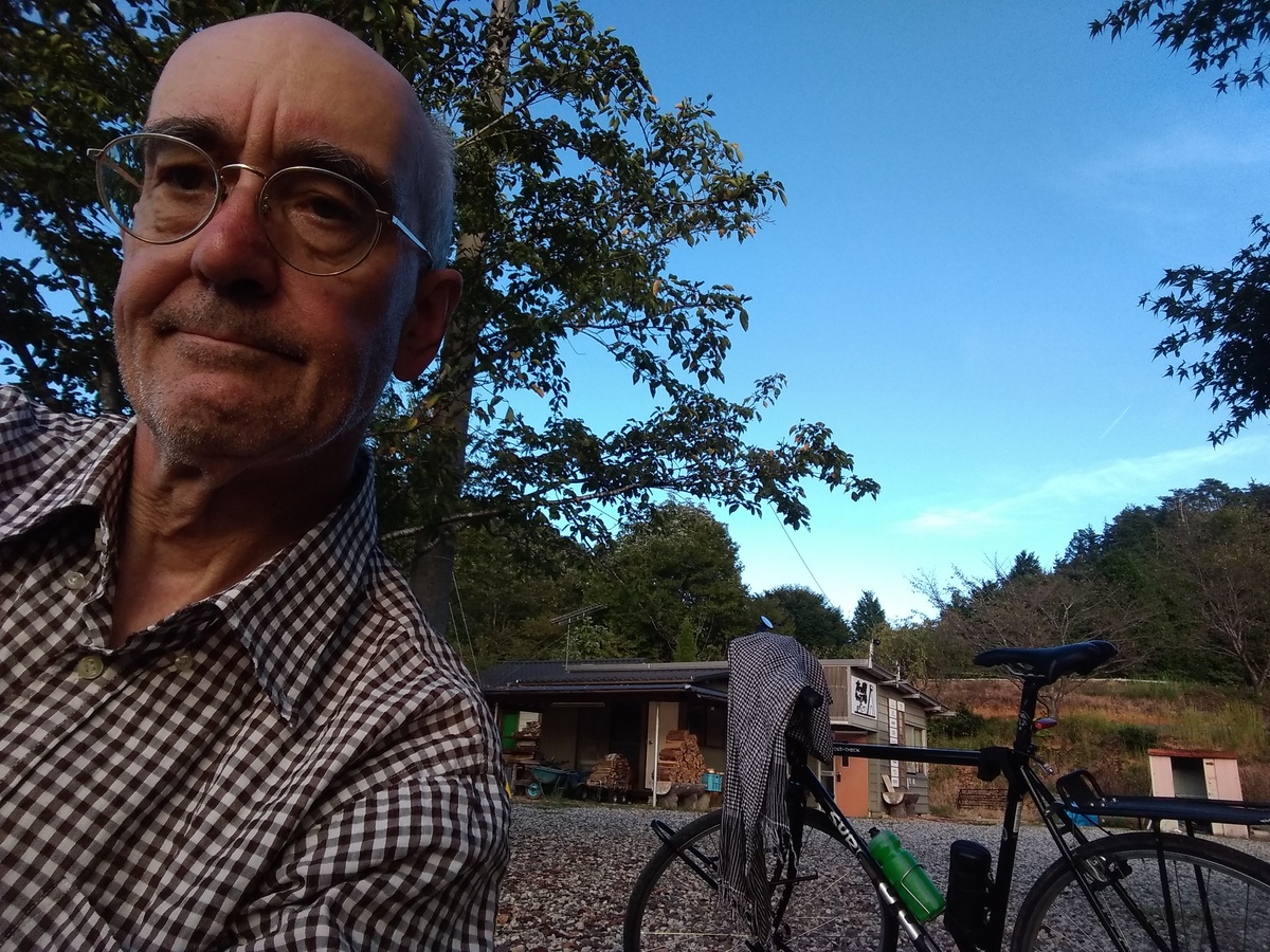 A chest-up photo of a bald man in a chequered shirt, with a tree shading a utility shed in the background, and a patch of blue sky beyond.