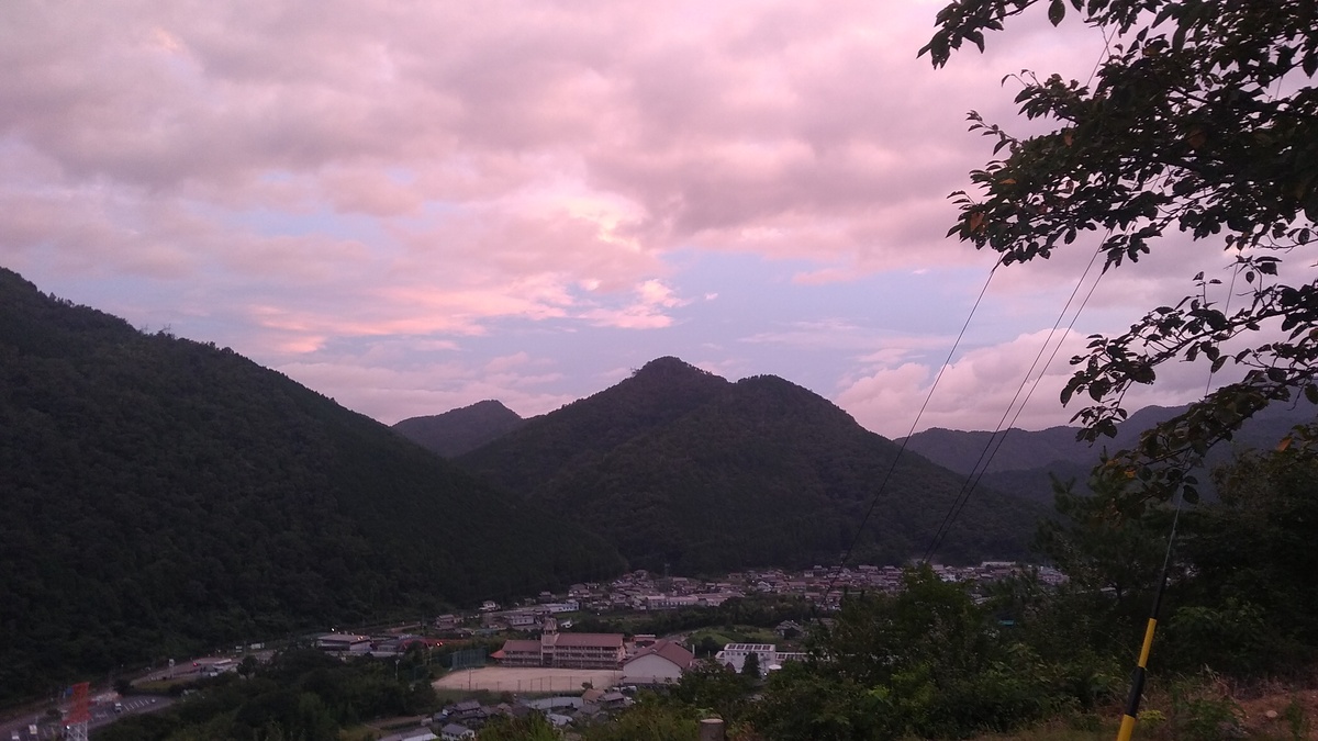 Blue sky over mountain tops, rimmed with clouds tinged with pink overhead. A village spreads out in the valley below.