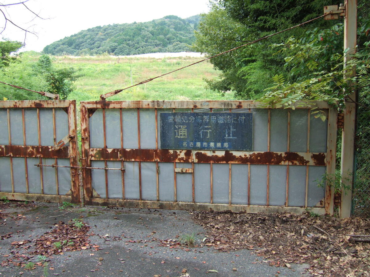 A rusty steel gate overgrown with brush bearing a sign reading (in English translation); “Roadway for the exclusive use of Aichi-Gifu Landfill: Entry is Prohibited”