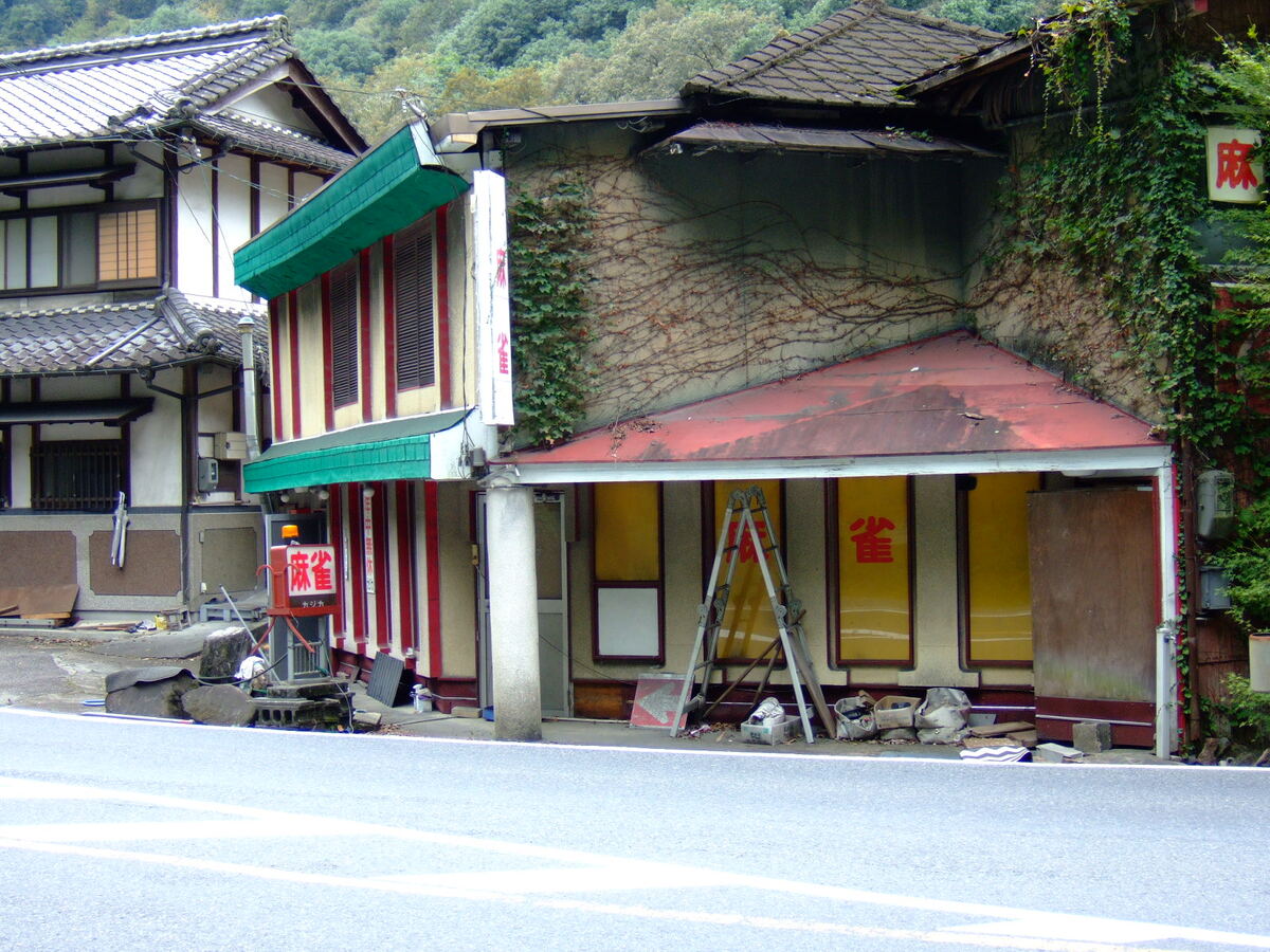 A two-story building with slate roof and vines adorning the face of the second-story wall, with green and red bunting over the entrance, and a sign reading (in English translation) “Mah-Jong.”