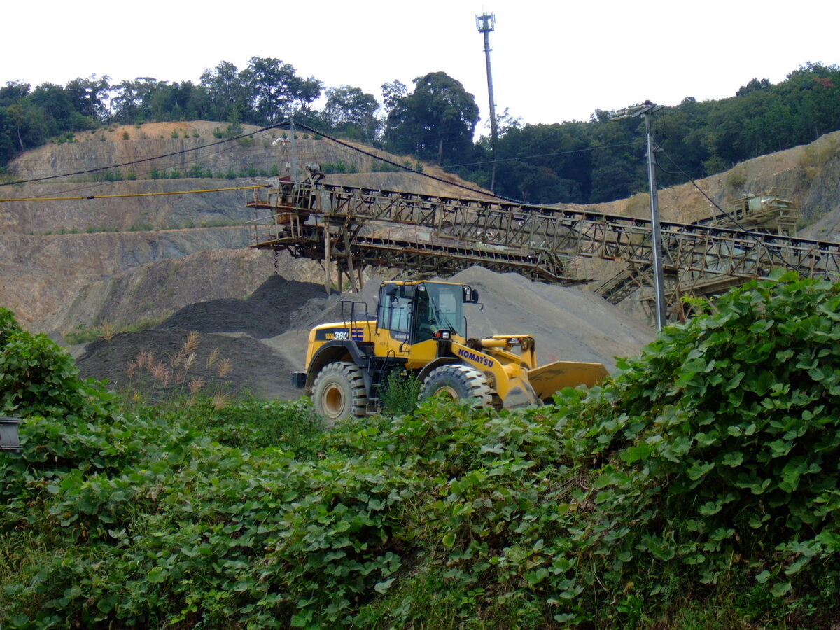 A massive skip-loader with an industrial conveyor in the background, standing on a hillside razed as part of a gravel pit. A G5 cell tower stands far in the distance higher up the hill.