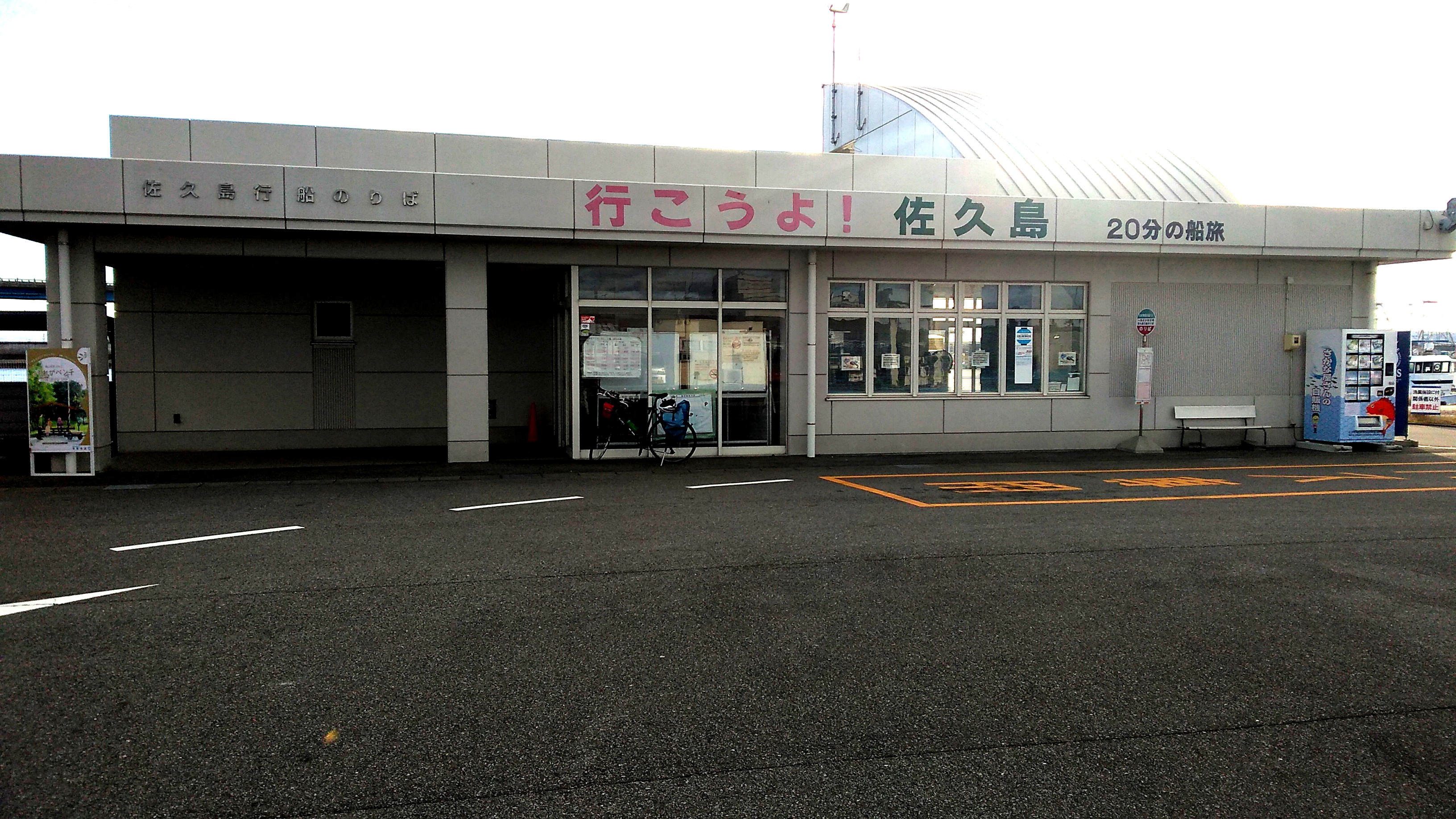 A grey one-story flat-roofed building with lettering on the side reading (in English translation) “Let’s Go! / Sakushima / a 20-minute ferry ride”