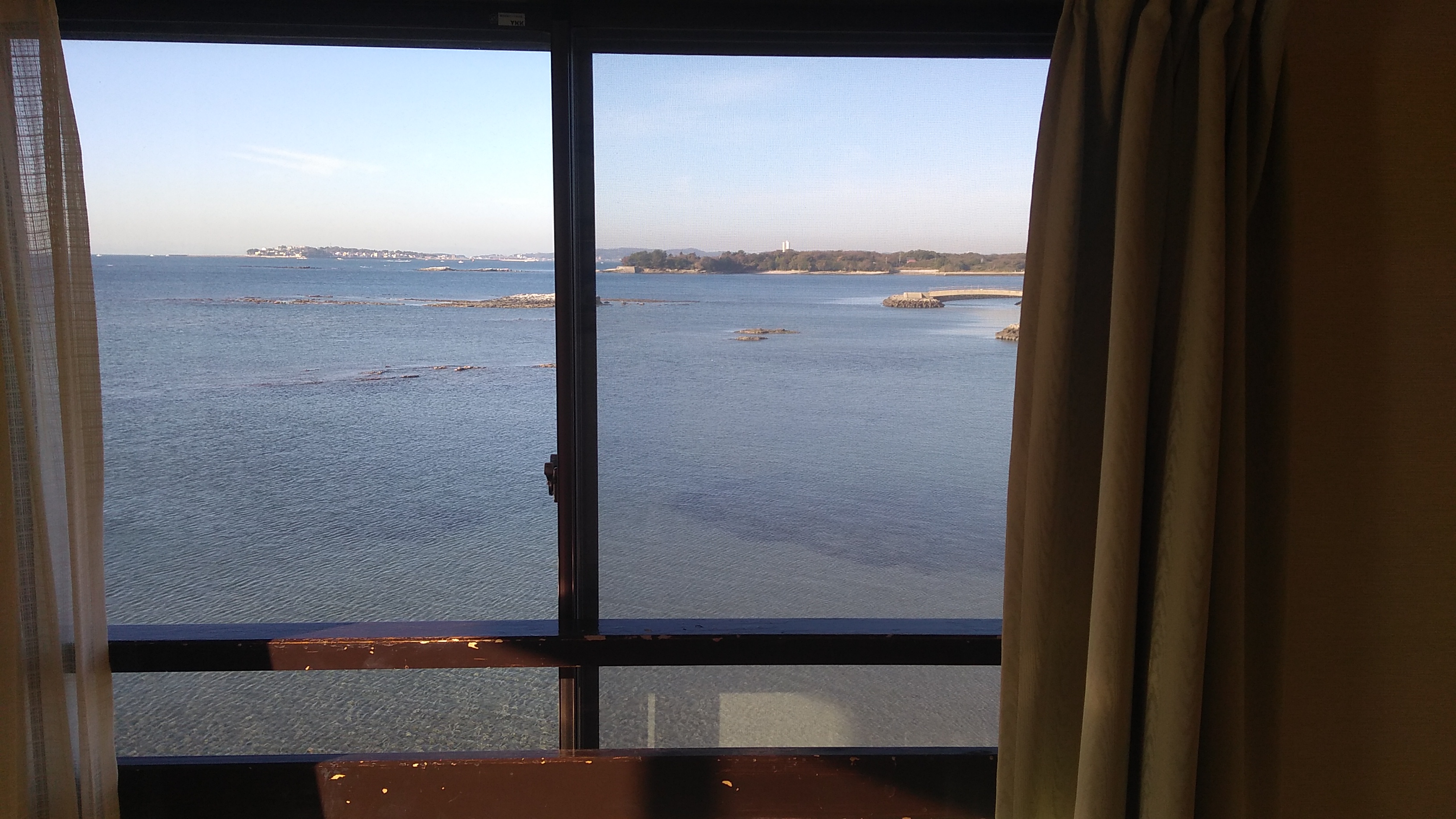 View through a second story window framed by beige curtains of a blue expanse of ocean water with a low-lying forested land mass in the distance.