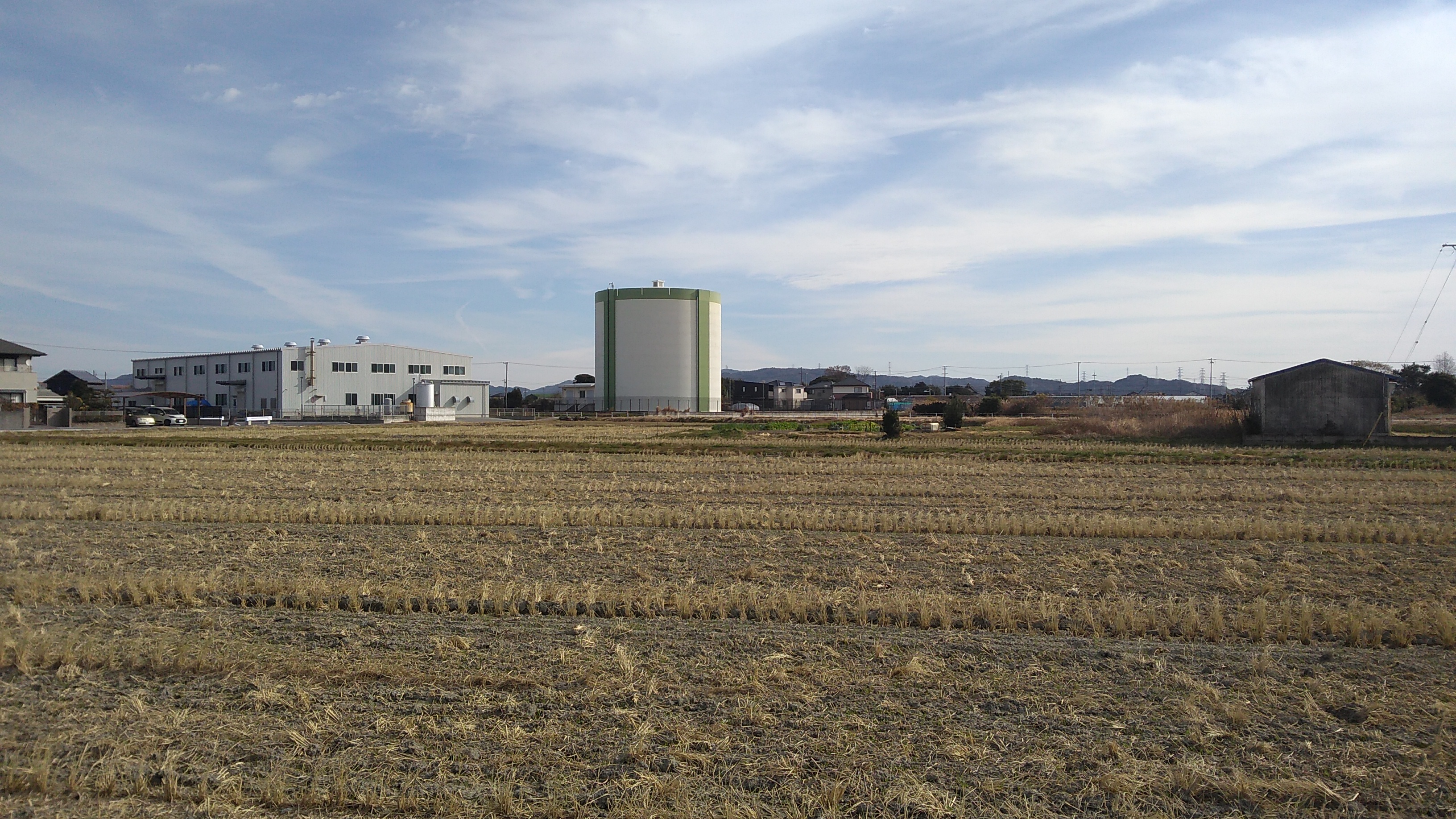 A squat tower that might be an agricultural silo or an industrial enclosure standing beyond an expanse of try stubble field under a blue sky with whispy clouds.