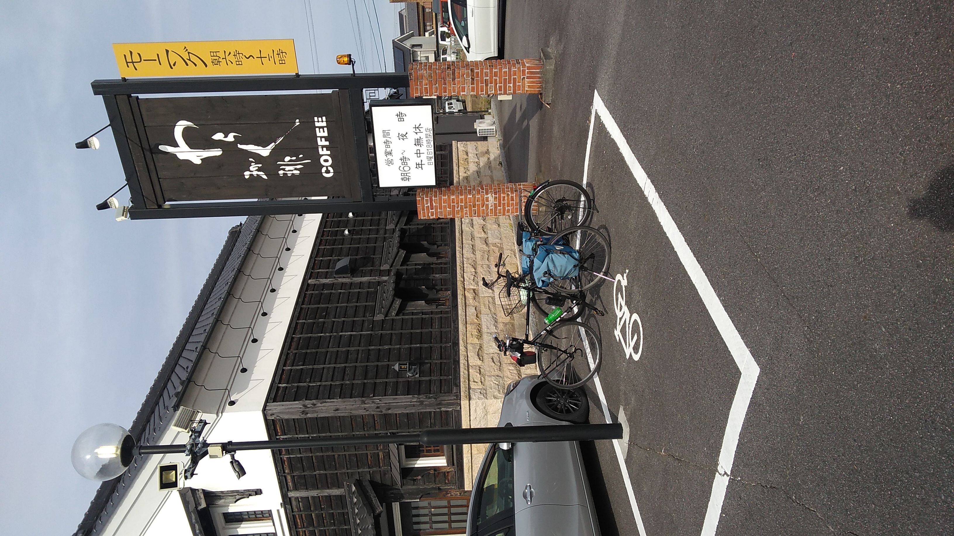 A black bicycle with blue panniers parked in front of coffee shop with a sign reading (in English translation): Waraku Coffee / Morning service 6:00–13:00 / Open hours 6:00–evening / All year / Sunday closing time 18:00