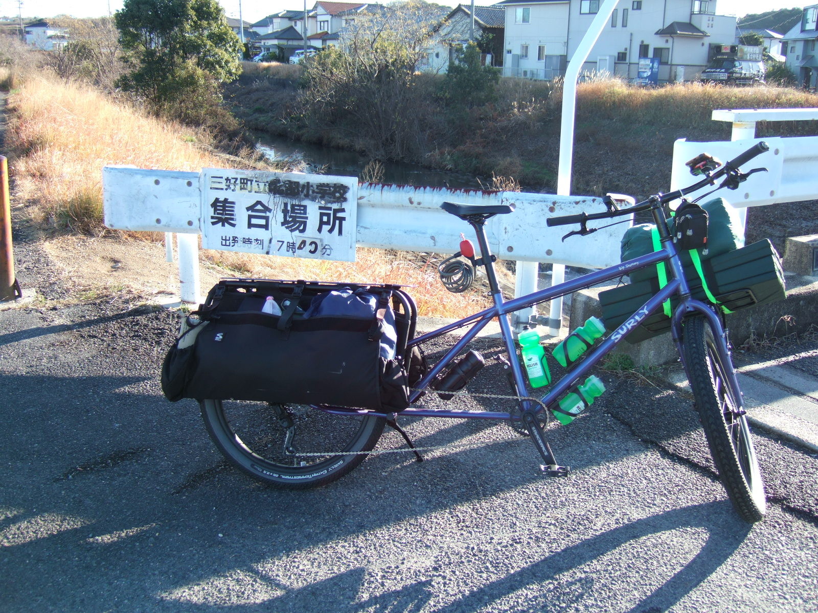 A loaded cargo bicycle parked at a bridge over an earthen canal with trees and dried grass on the banks. A sign on a guardrail behind the bike is partly obscured by black spray paint over the school name, but seems to read “Miyoshi North District County Elementary School/Gathering Point/Departure Time 7:40” (三好町立北部小学校・集合場所・出発時間７時４０分). Not sure what the reason for the splotch of black paint is, but the sign must be at least 13 years old, since Miyoshi County became Miyoshi City in 2010.