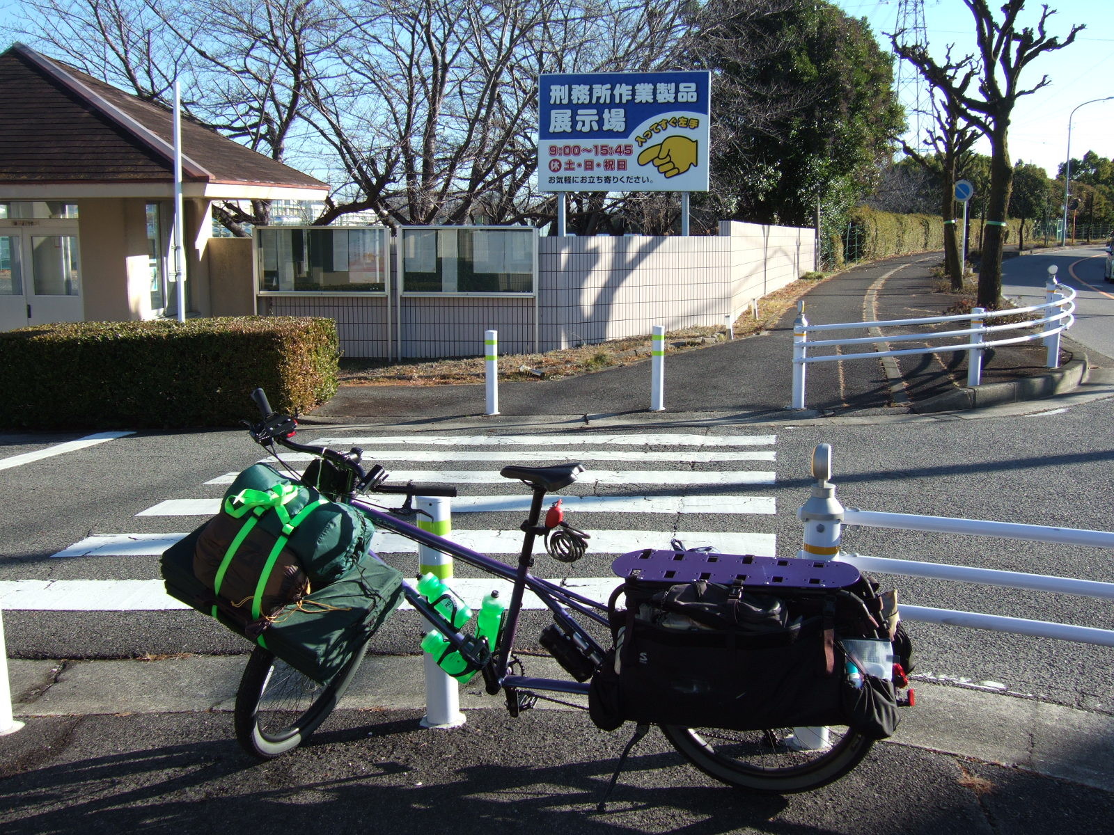 A loaded cargo bicycle parked at a zebra crossing spanning the entrance to the grounds of a prison. On the far side of the zebra crossing is a sign reading “Exhibition Gallery of the Products of Prison Labor/Open 9:00-15:45, except Saturdays, Sundays, and Holidays/Immediately to the left of the entrance/Visitors welcome” (刑務所作業製品展示場・９：００−１５：４５㊡土・日・祝日・入ってすぐ左手・お気軽にお立ち寄りください). A cartoon finger points to the opening hours.