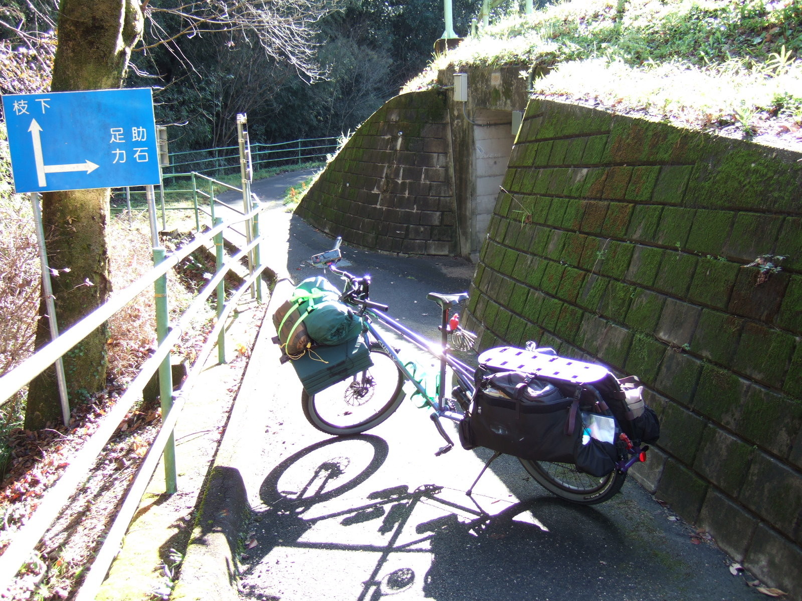 A loaded cargo bike parked in a narrow paved pathway, with a steep stonework embankment on its right. The entrance to a tunnel under the embankment is to the right, and there is a sign to the left of the path with arrows indicating destinations Sanage (猿投) straight on, and Asuke (足助) and Chikaraishi (力石) to the right through the tunnel.