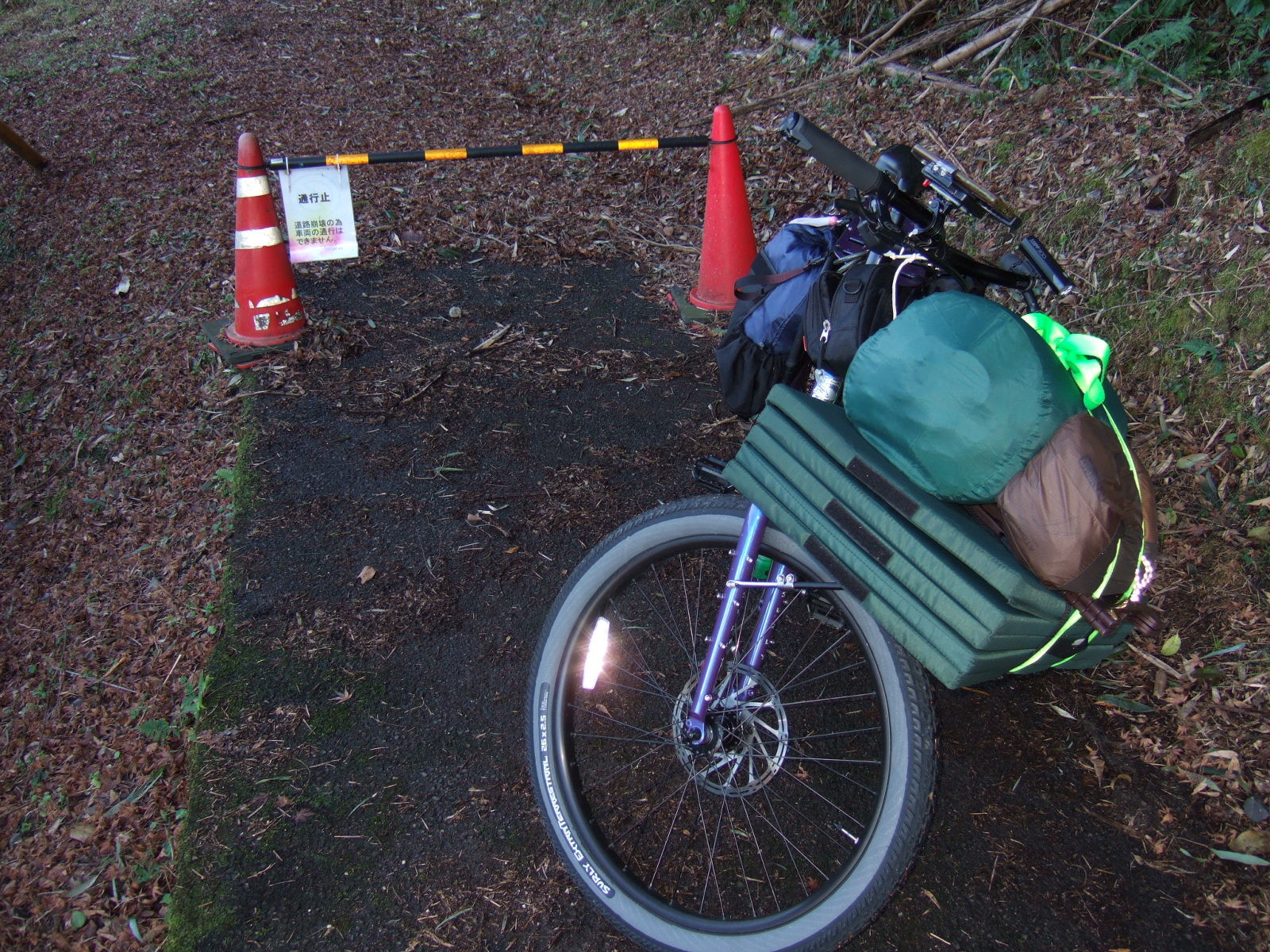 A loaded cargo bike parked on a forest floor, with traffic cones behind supporting a plastic rail on which is hung a sign reading: “No Through Road/Due to destruction of the road, vehicles cannot pass” (通行止・道路崩壊の為車両の通行はできません).
