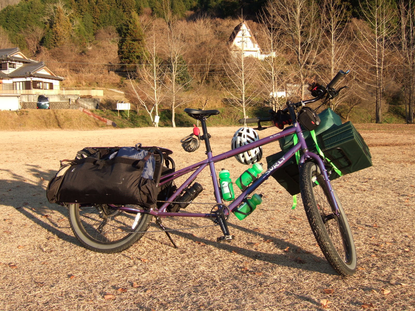 A loaded cargo bike parked on a field of close-dropped dried grass, with an A-frame house on a wooded hill in the background.