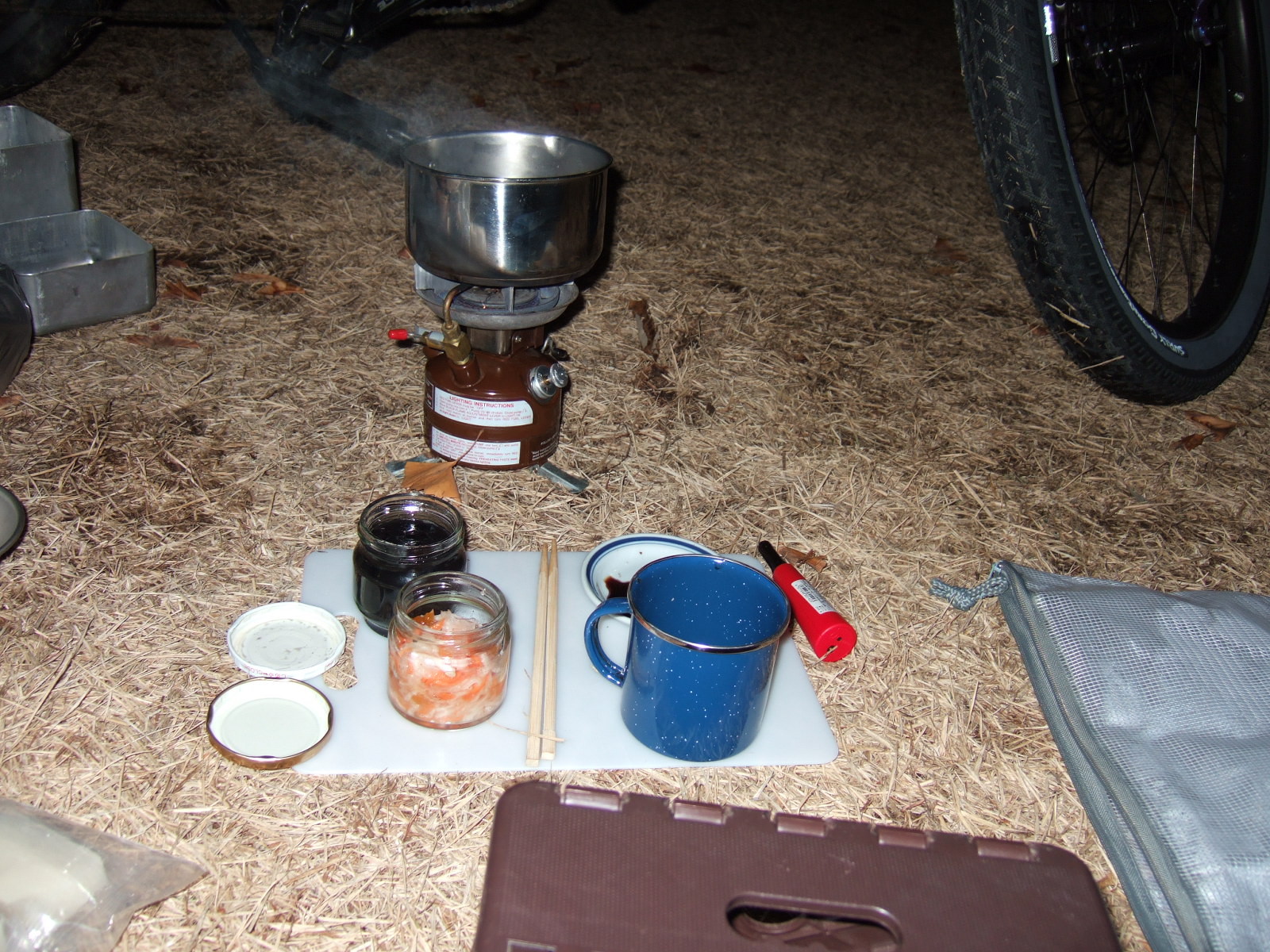 A small jar of sweet-and-sour carrot and radish salad, a small jar sweet black beans, a coffee cup and small plate of soy sauce on a plastic cutting board, resting on the ground. Beyond the cutting board a pot of water is boiling on a small gasoline camp stove.