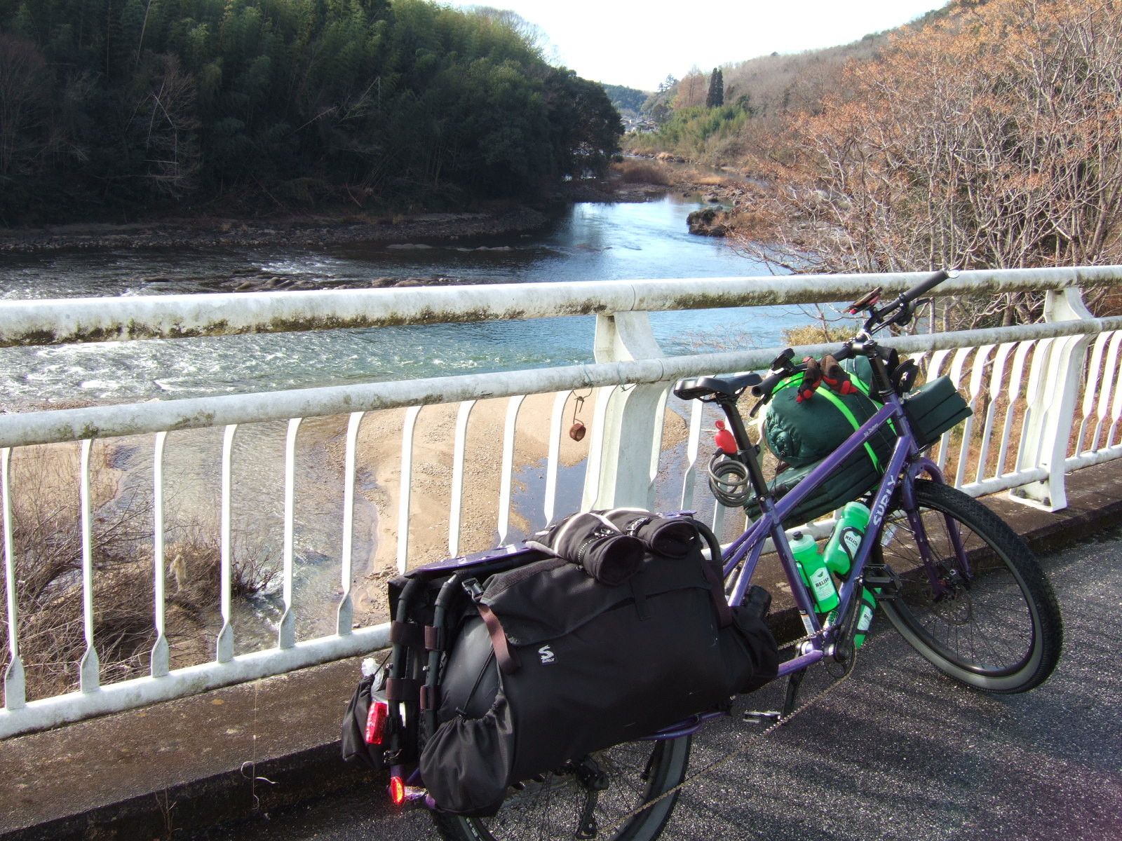 A loaded cargo bike parked at a painted steel railing, overlooking the view of a river winding into the distance between wooded banks, with a small village visible very far down at a distant bend in the river.
