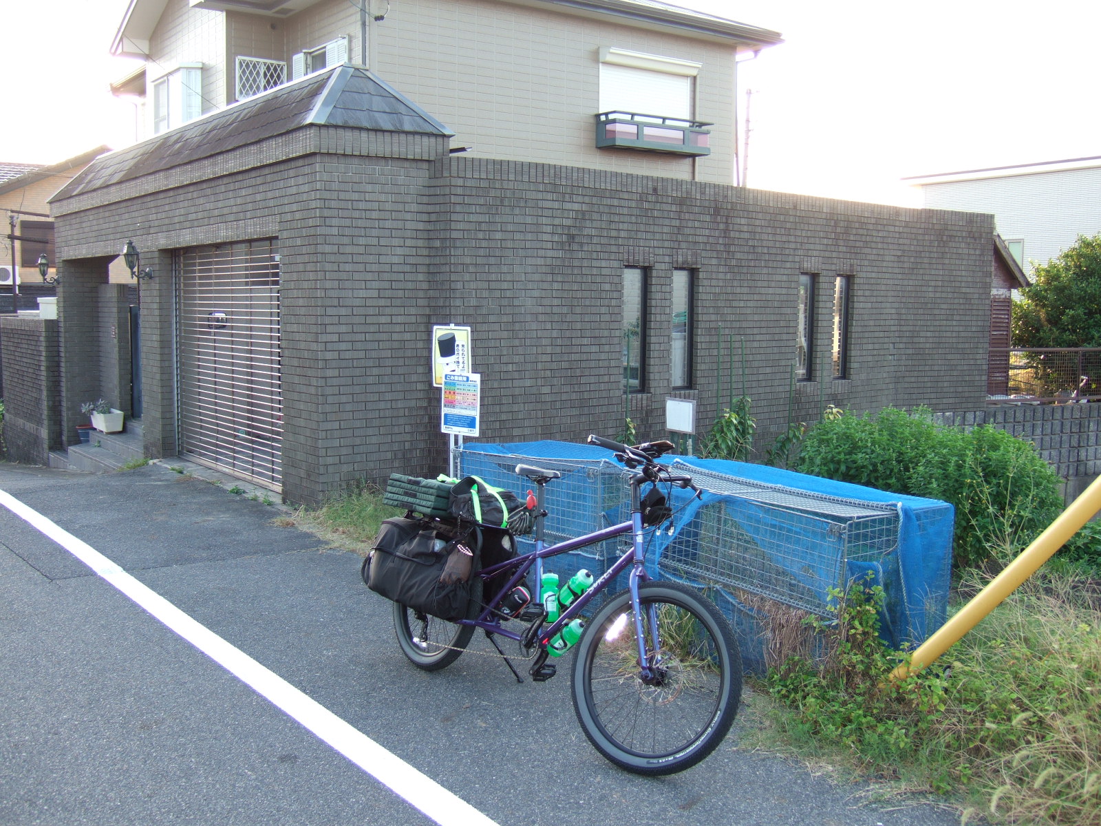 A Surly Big Dummy cargo bike parked at the side of a road, with two steel cages sheathed in blue netting behind, and house enclosed in a curtilage wall behind.