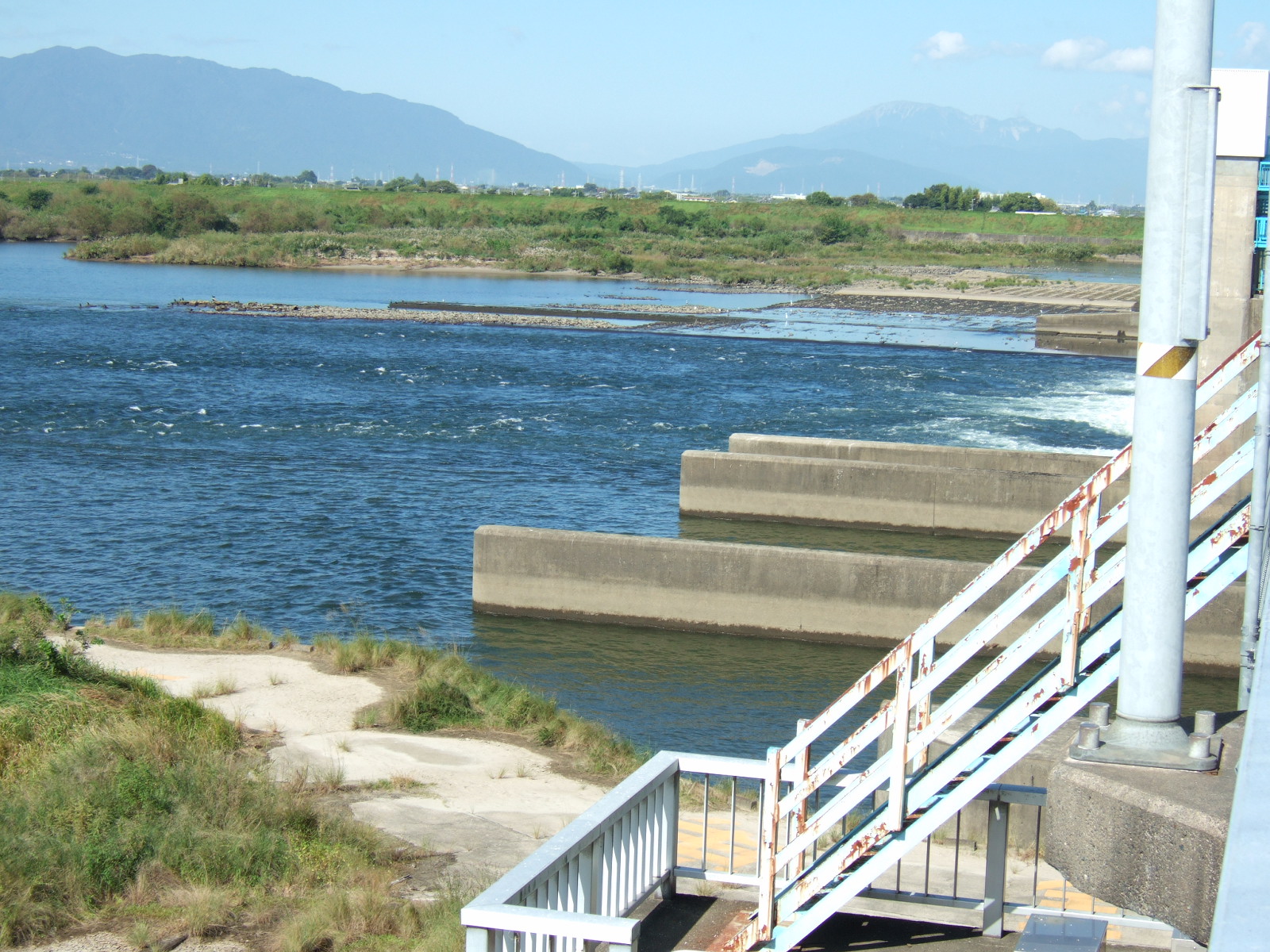 A expanse of clear blue water lies below the dam in a wide river. Mountains on the horizon are shrouded in mist.