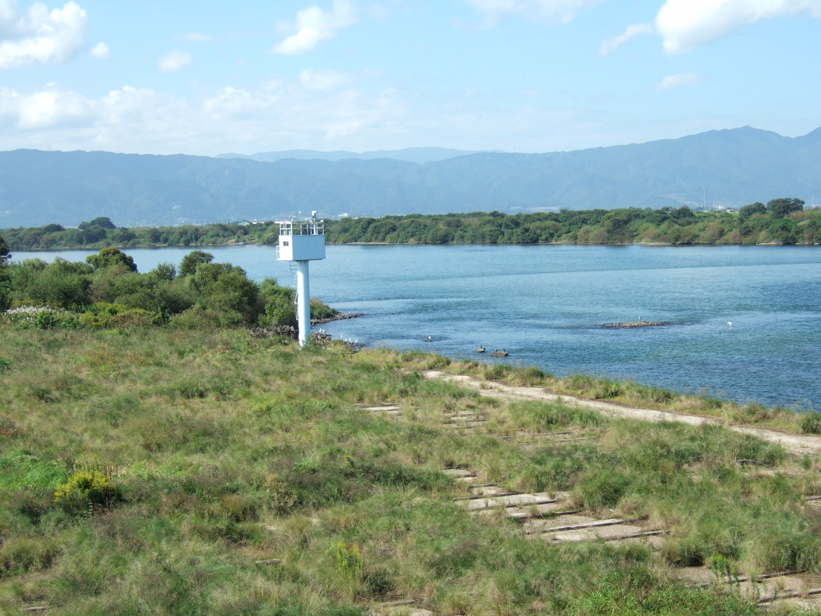 A grassy shoreline borders a broad river. A single tower (a monitoring station?) stands at the water’s edge. A mountain range in the distance is shrouded in mist.