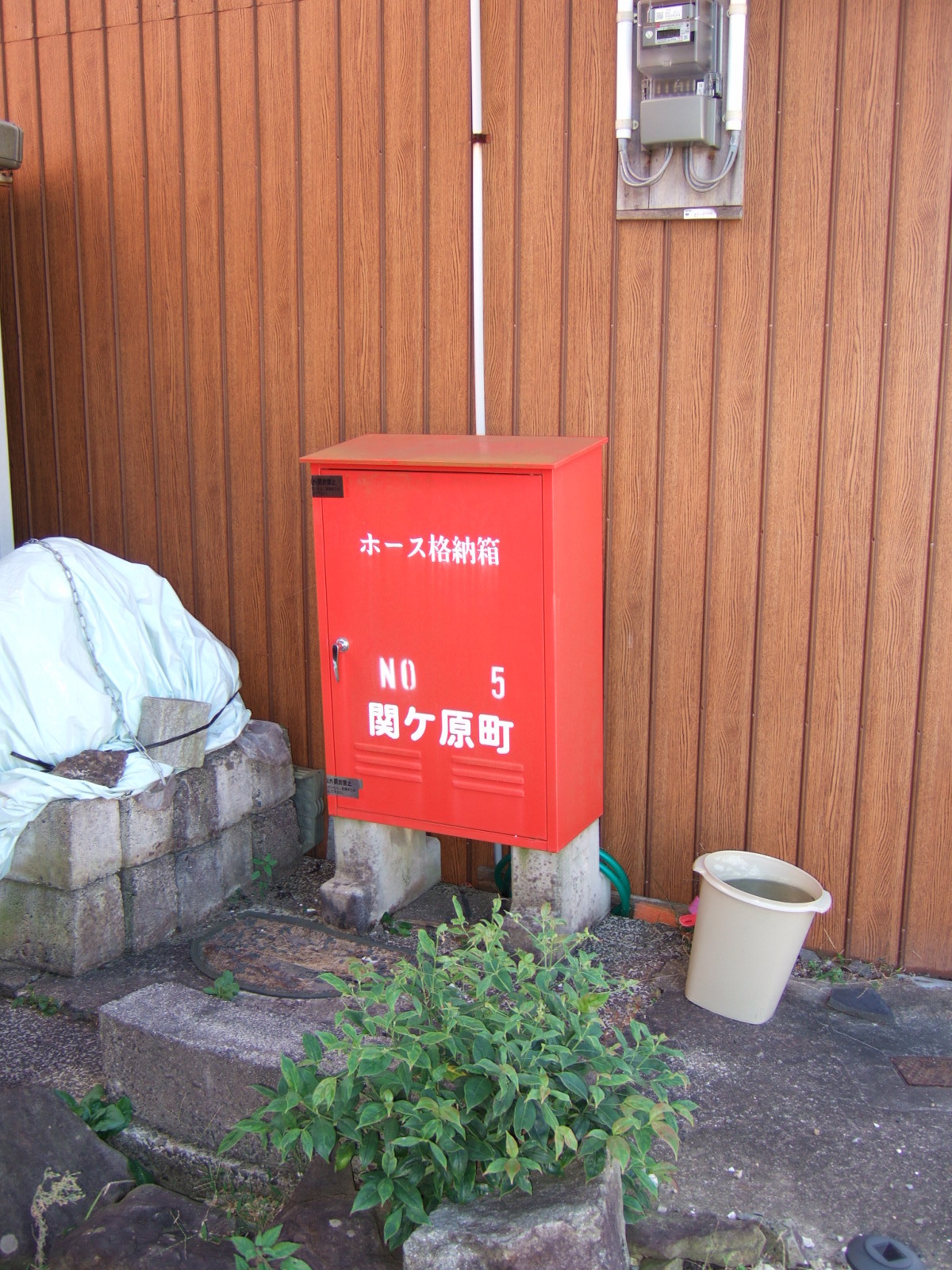 A tin box painted red, standing on two cinder blocks against the aluminum siding of a building. An electric meter is fastened to the outside of the building. Lettering on the box reads: Hose Container/Number 5/Sekigahara County\nホース格納箱・NO 5・関ヶ原町