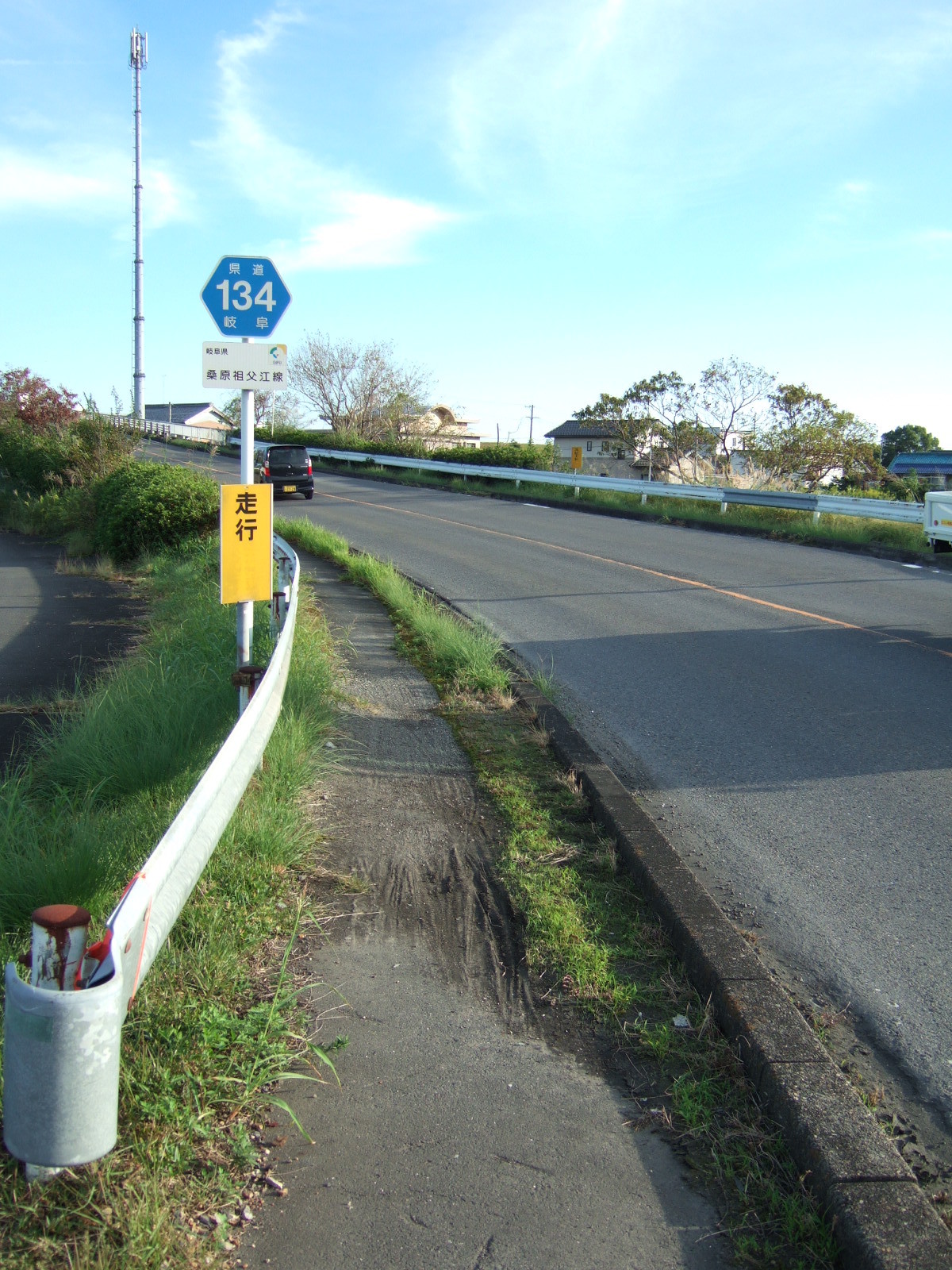 A narrow path with a guard rail to the left and a low concrete curb to the right lies next to a trafficked road stretching upward toward a bridge crossing. A sign indicates this is Route 134, and a yellow sign below tersely marks this as “walking path” 歩行