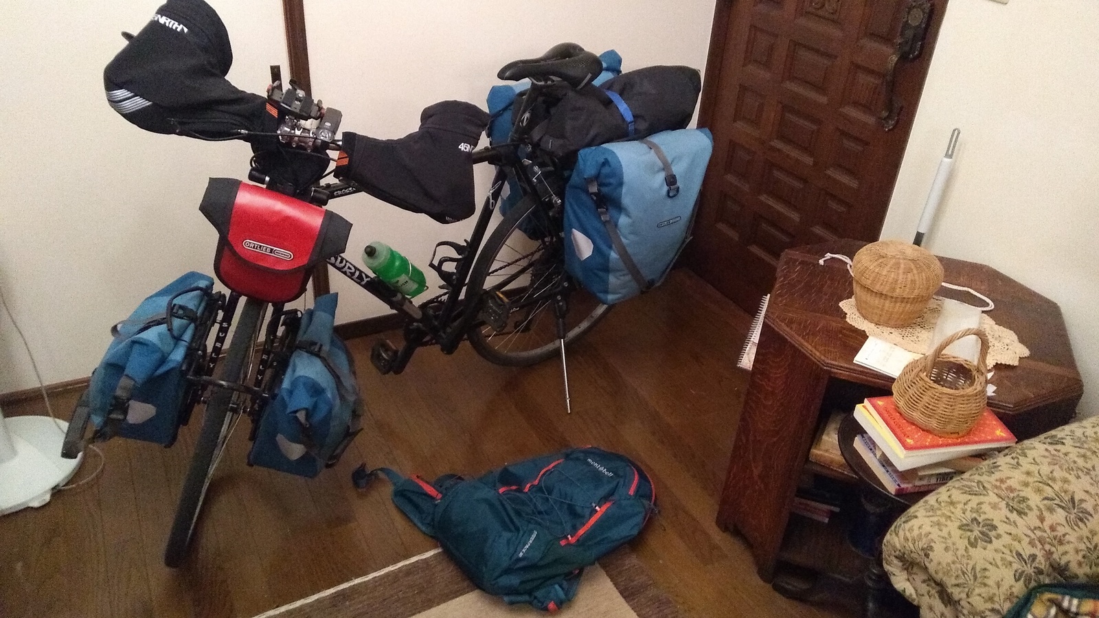 A black bicycle with a small red handlebar bag, bar mitts, and blue panniers front and rear, parked in the corner of a living room with a day pack lying on the floor in front of the bike.