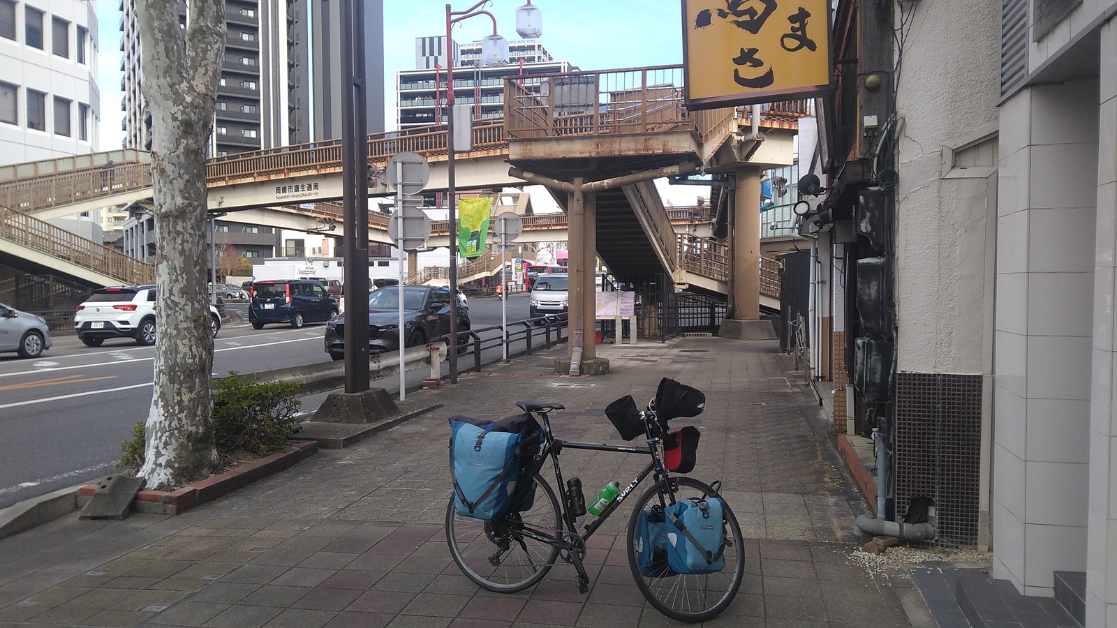 A bicycle parked on a city sidewalk, with a rusting steel overcrossing and stairs above an intersection in the background.