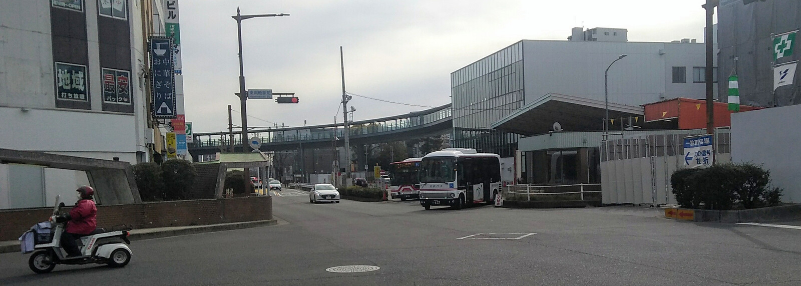 A large empty intersection paved in grey asphalt under a grey sky with square grey buildings to the left and right. A long enclosed overhead walkway is visible in the distance beyond the intersection.