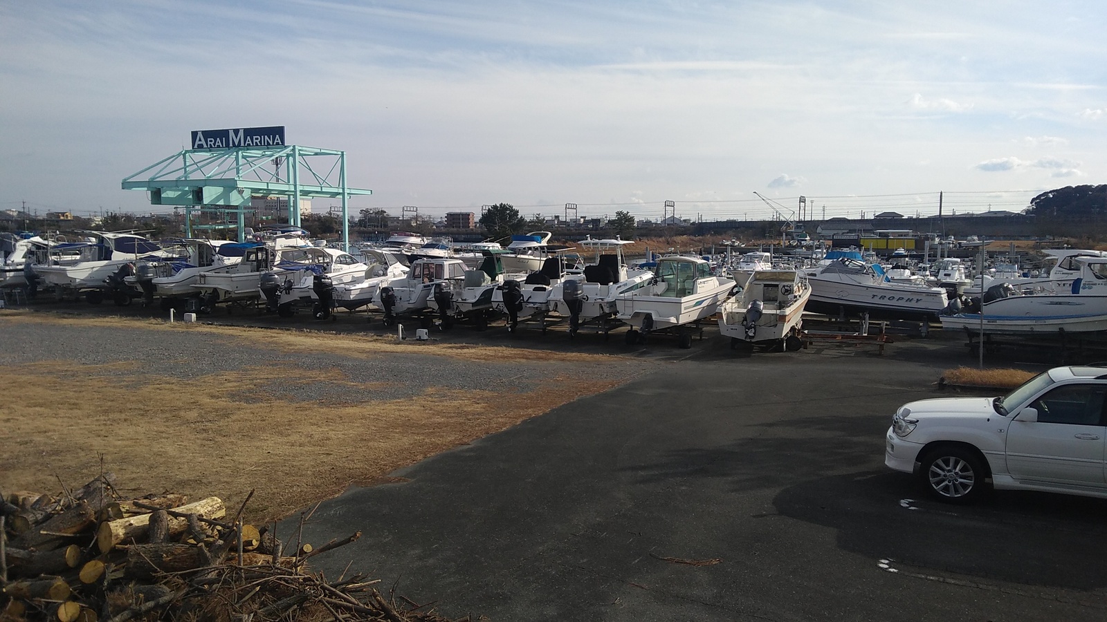 A cluster of perhaps two dozen motorboats are parked in a lot beside the road, with a blue-green steel latticework in their midst bearing a sign reading “Arai Marina”