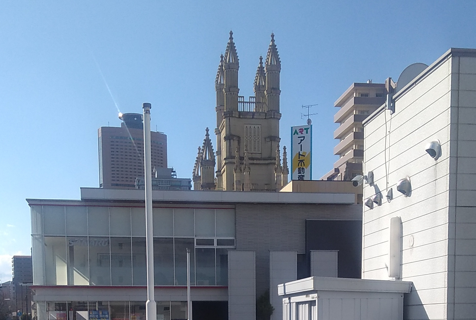 Four tall spires and several lesser ones in the style reminiscent of of Antoni Gaudi rise into a blue sky beyond a nondescript utilitarian square building in the foreground.