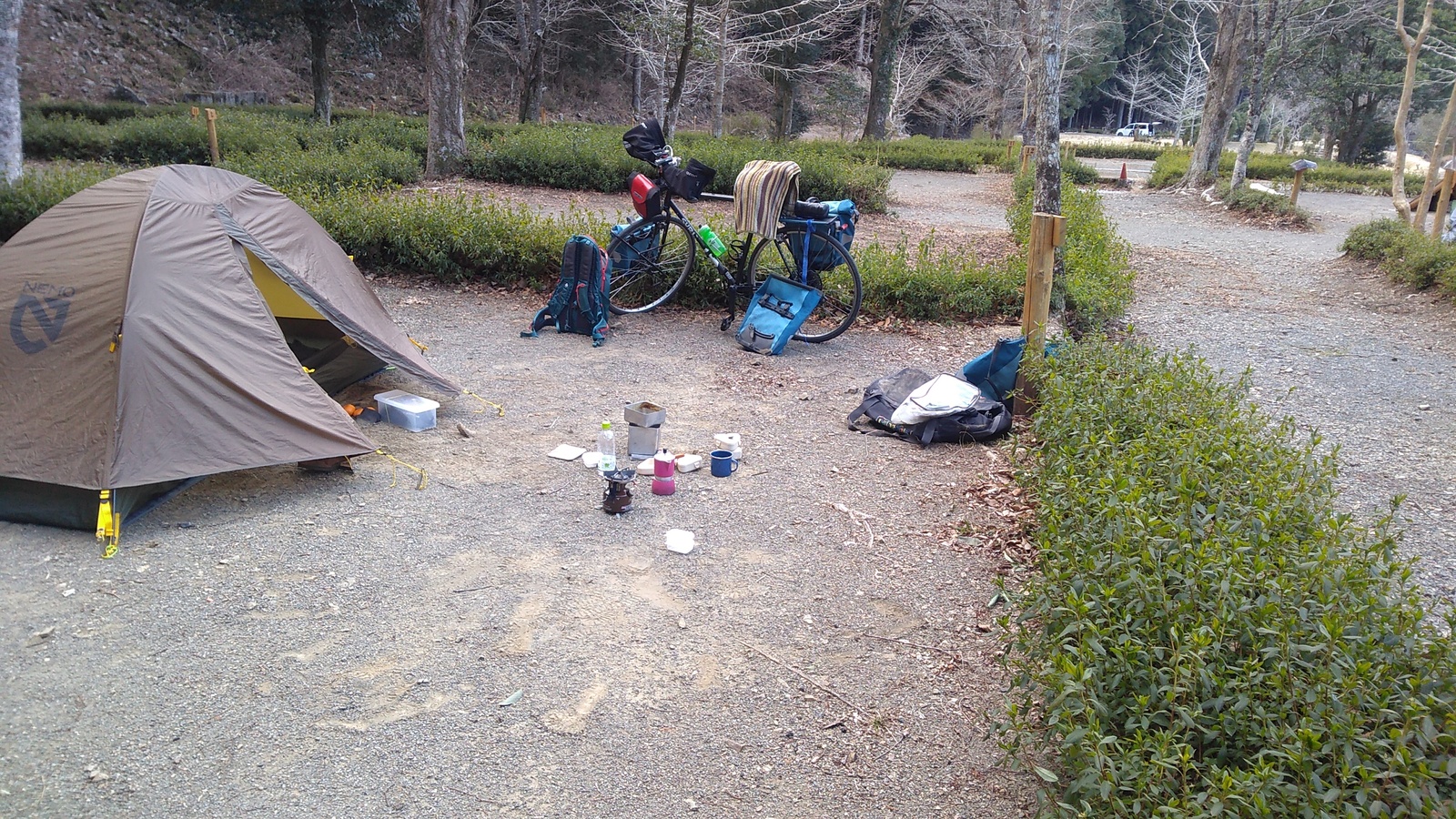 One of several individual campsites, each edged by low-lying hedges. In this one there is a one-man tent, and to its right a touring bicycle, a pile of assorted luggage, and with a camp stove with a coffee pot in the middle of the scene.