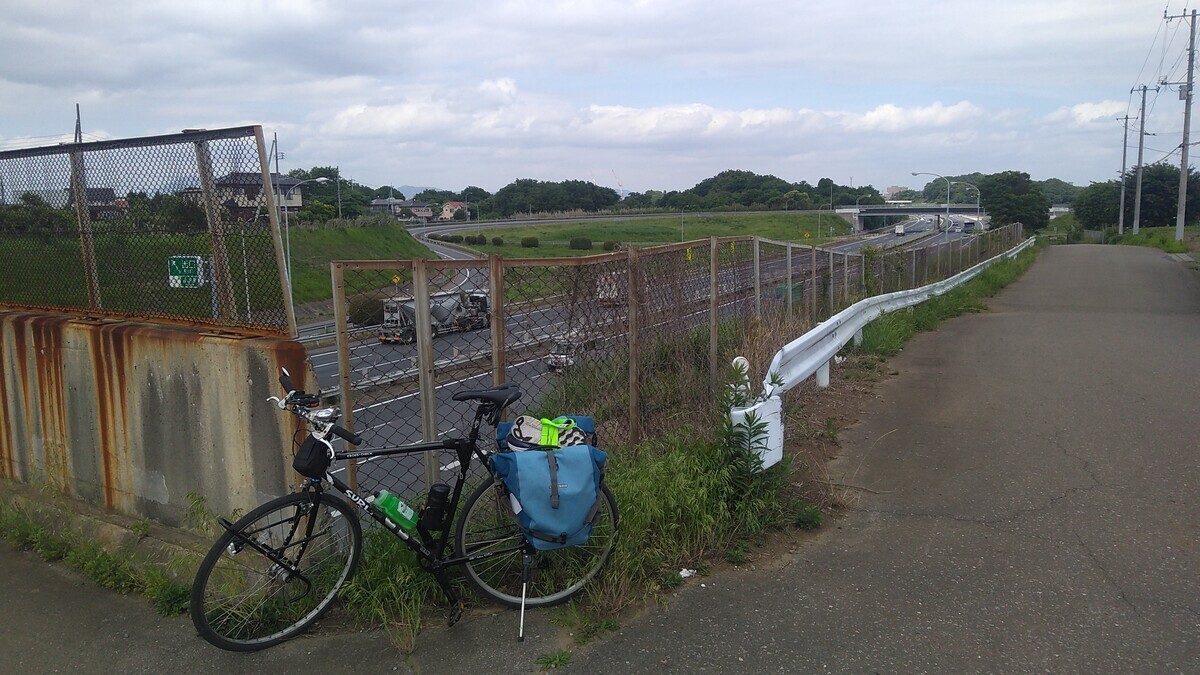 A black bicycle with blue panniers parked at the overlook of a freeway cutting, at the entry point to an overpass. A rust-stained concrete barrier is behind the bicycle, and in the background a six-lane freeway is visible.