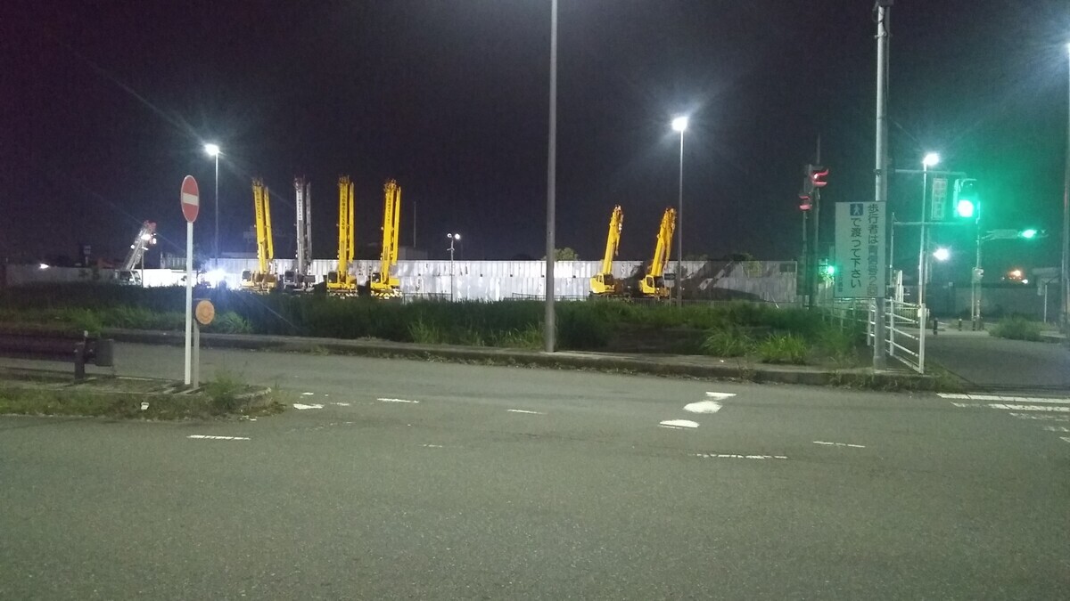 An evening view of seven large motorized cranes in a parking lot across the street  from the park,  brilliantly illuminated by halogen lamps.