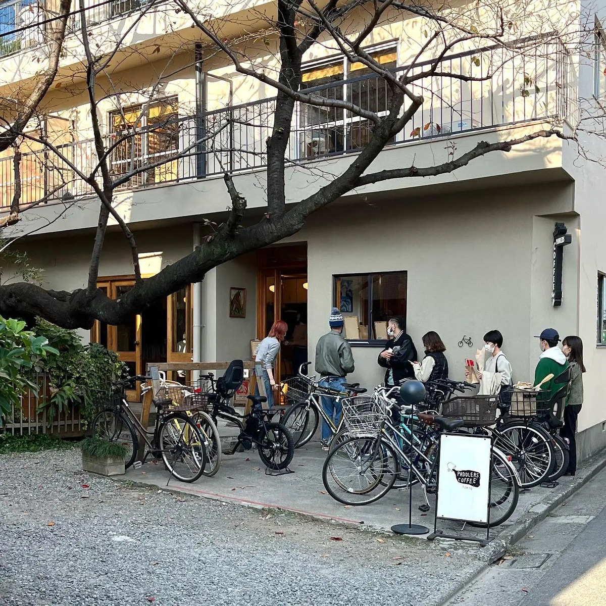 Entrance to a coffee shop, with a number of bicycles parked in front, and queue of customers awaiting entry. The building is a nondescript grey stucco, the entrance doorway is framed in varnished wood, and a large tree branch leans in across the scene.