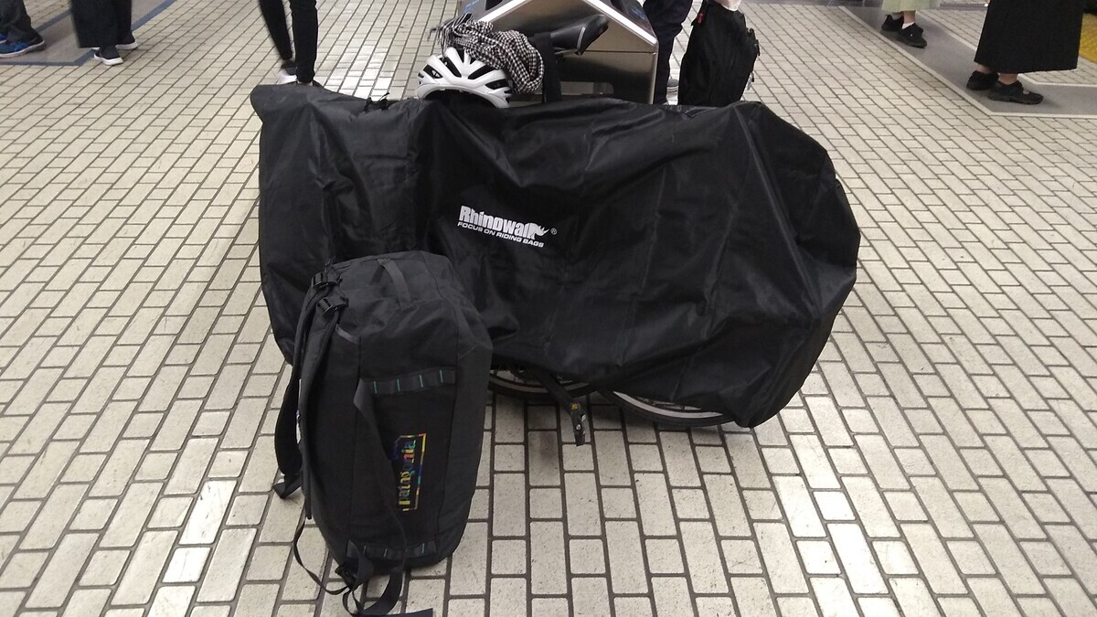 A bicycle wrapped in a carry bag and a 55-liter duffle bag standing in front of garbage bins on the Shinkansen platform at Shin-Yokohama station.