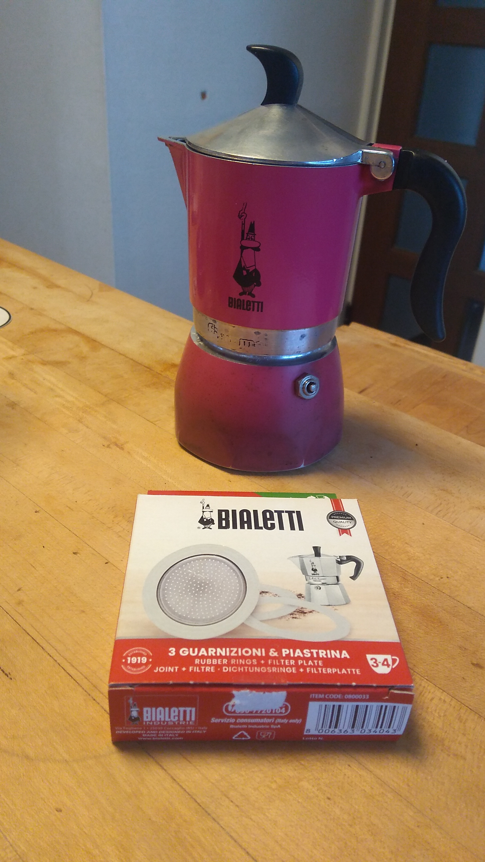 A fully assembled induction coffee maker, with the Bialetti logo of a mustached cartoon figure with one arm raised high with a pointed finger as if making an emphatic statement. A box of replacement seals sits in front of the assembled coffee maker.