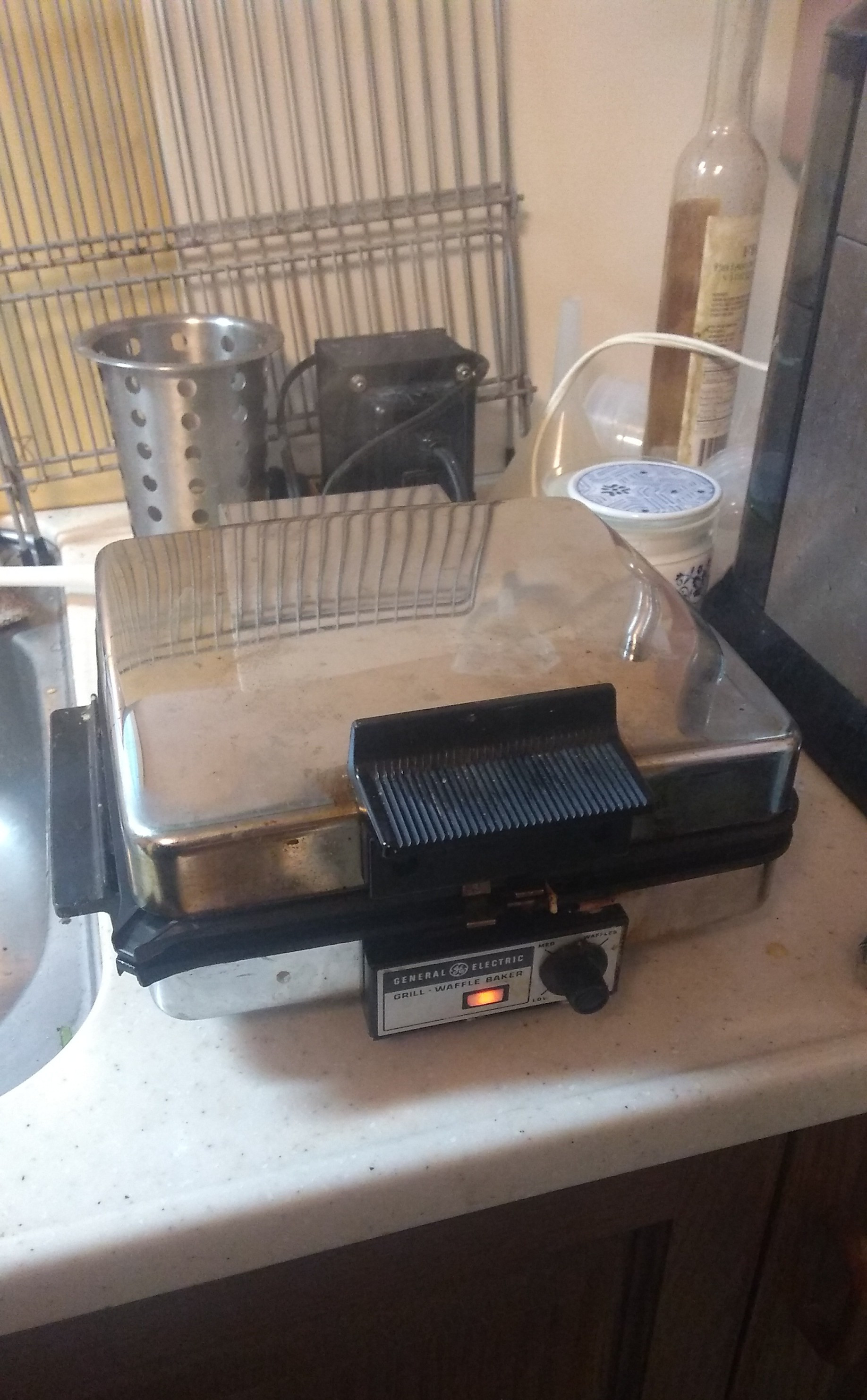 A stainless steel electric waffle iron by General Electric, sitting on a kitchen counter with the lid closed.